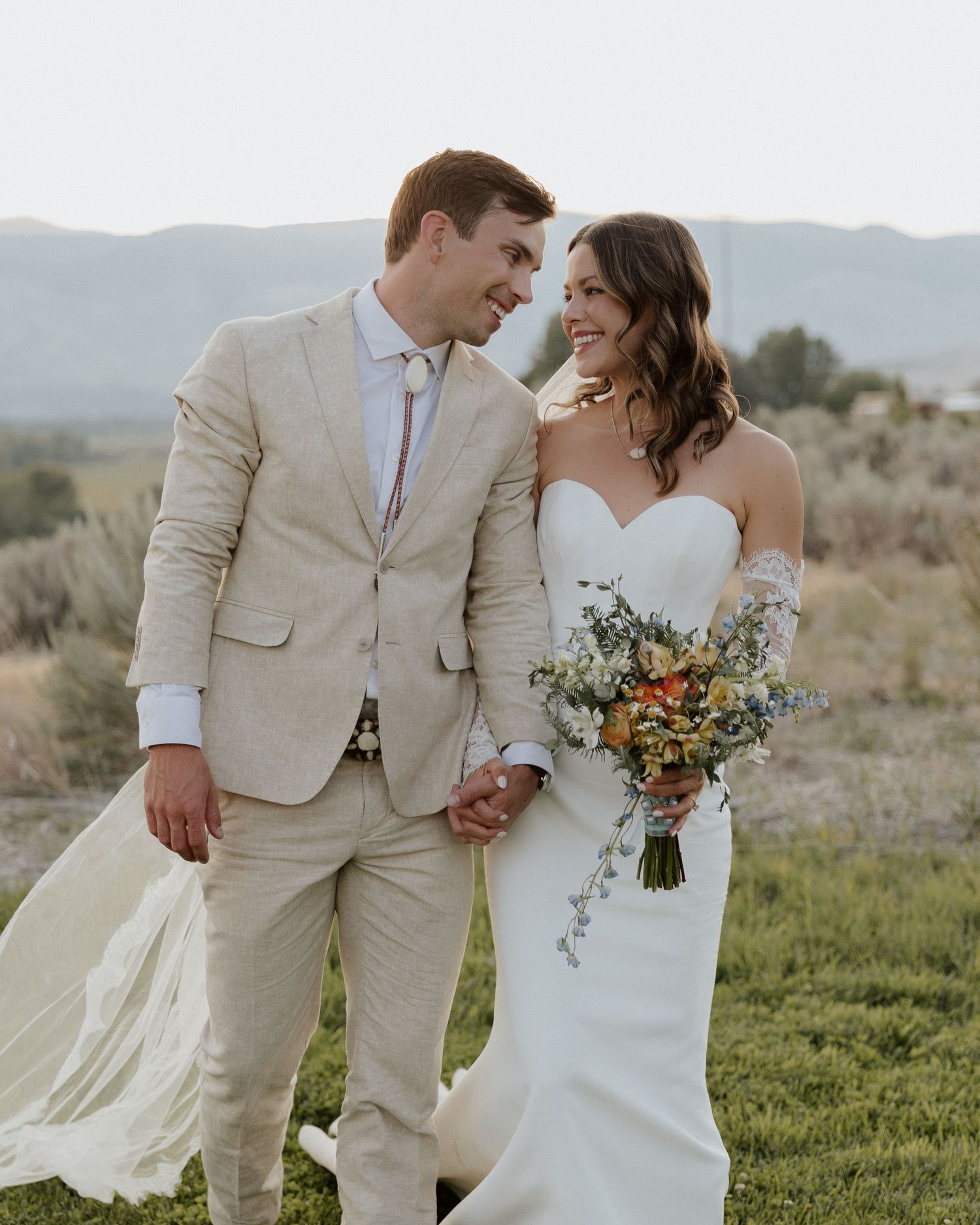 Wedding couple smiles together on grassy hillside with bride holding rustic bouquet.