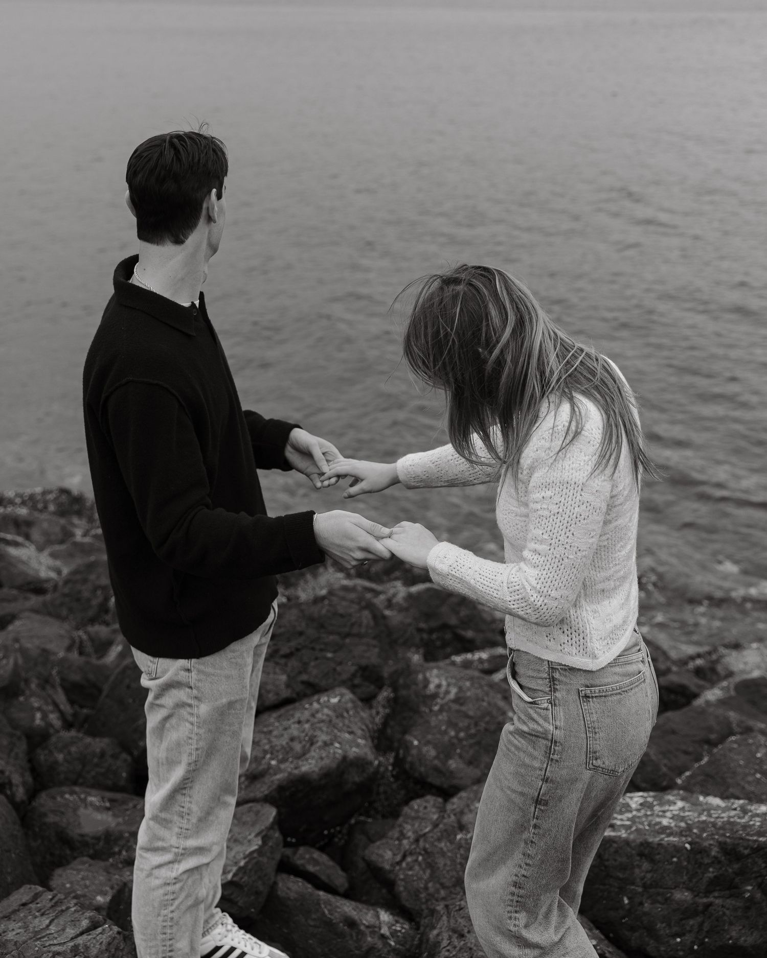 A couple shares a tender moment on rocky coastal cliffs overlooking a misty ocean in black and white.
