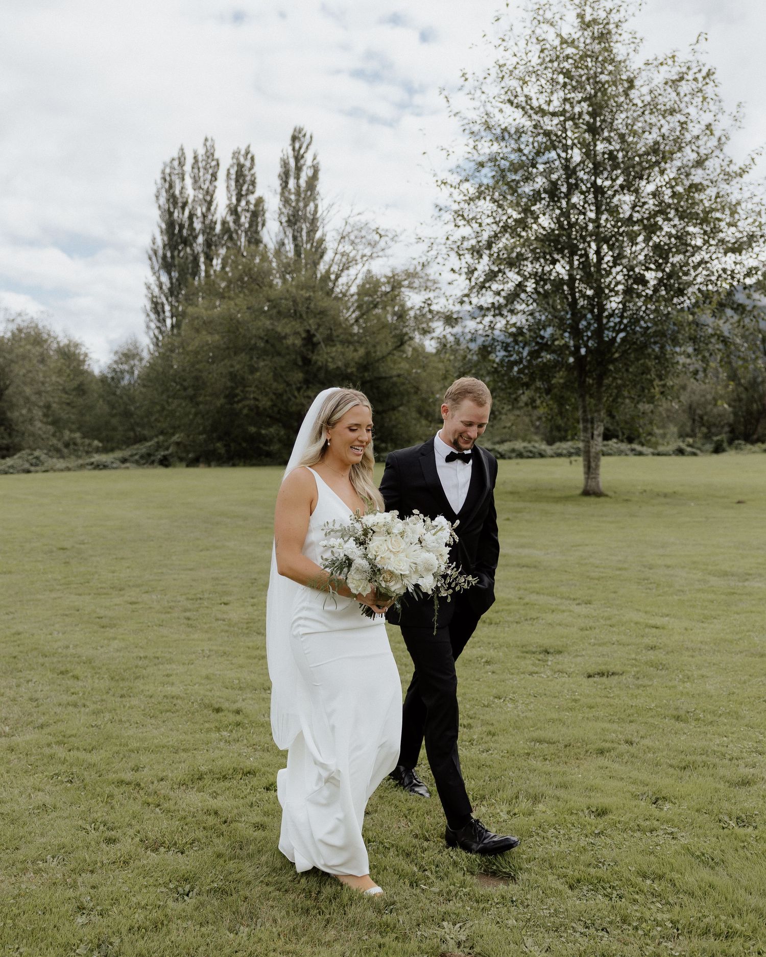Bride in flowing white dress walks with groom across green lawn during wedding.