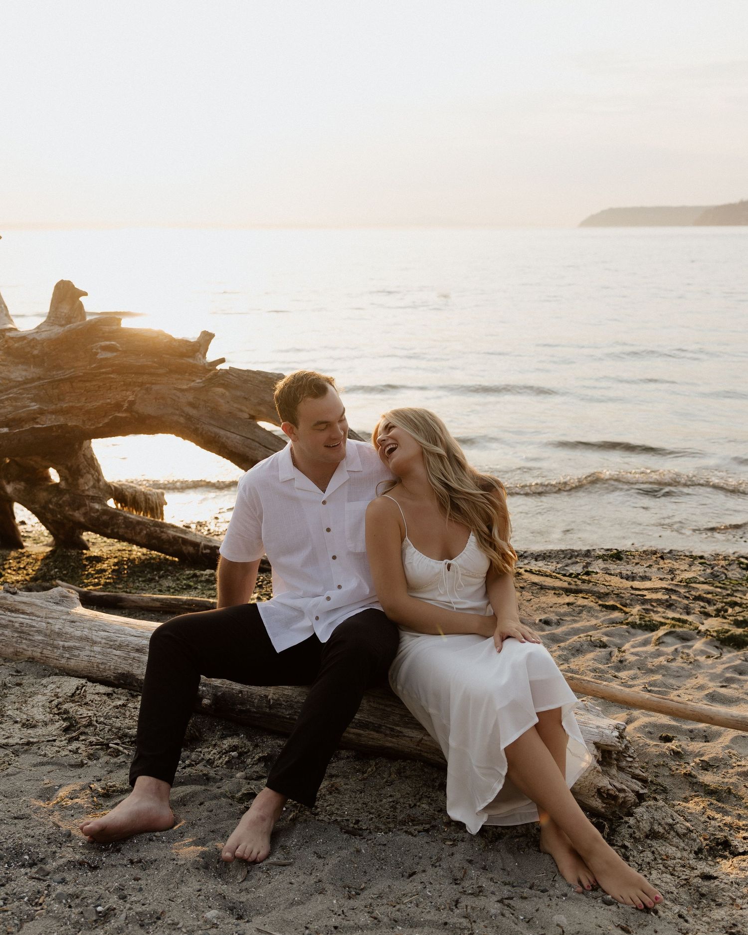 A romantic couple sits on driftwood at sunset on a beach wearing white and black attire.