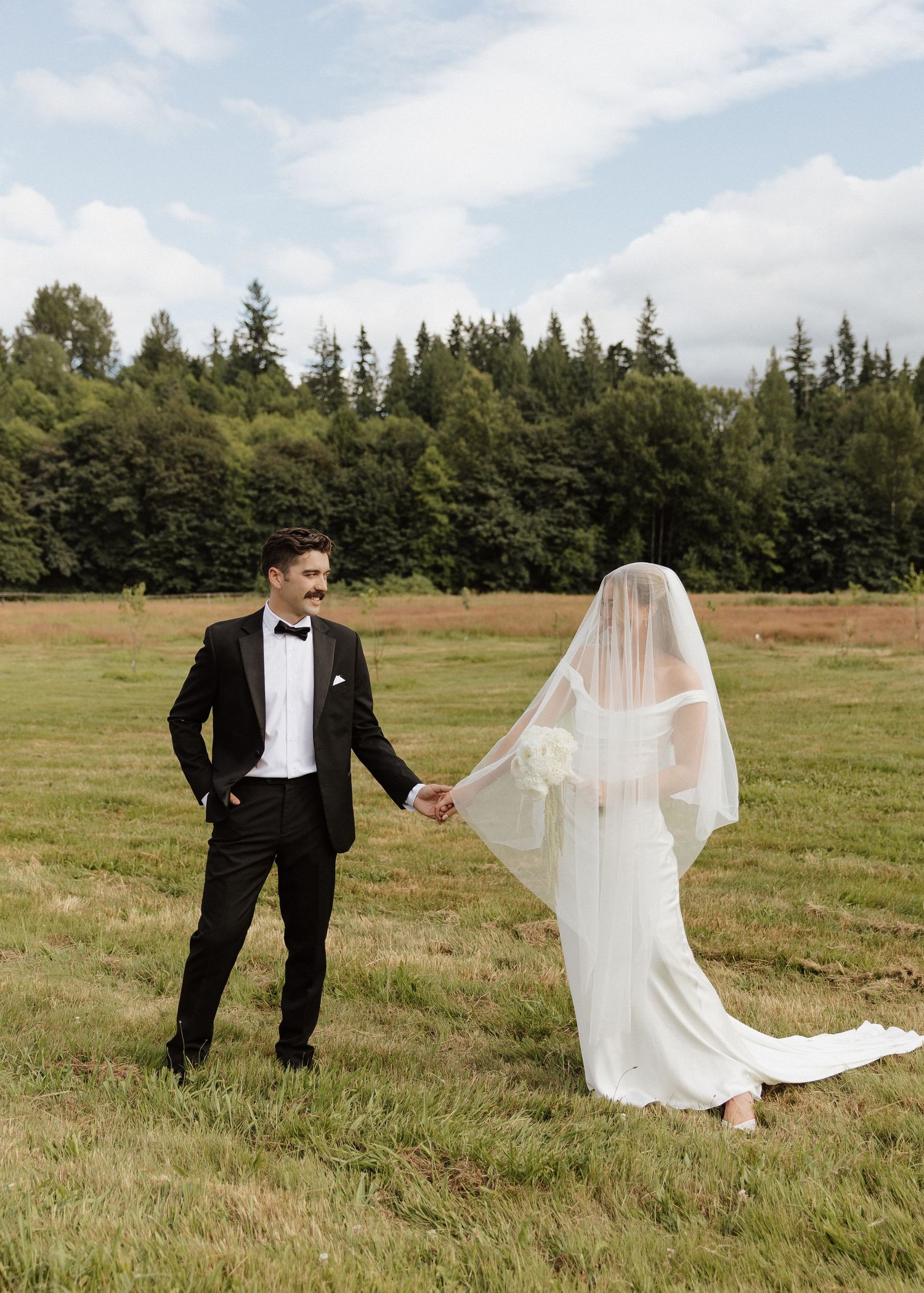 A bride and groom hold hands in a lush green field, she wears a flowing veil and he a classic black tuxedo.