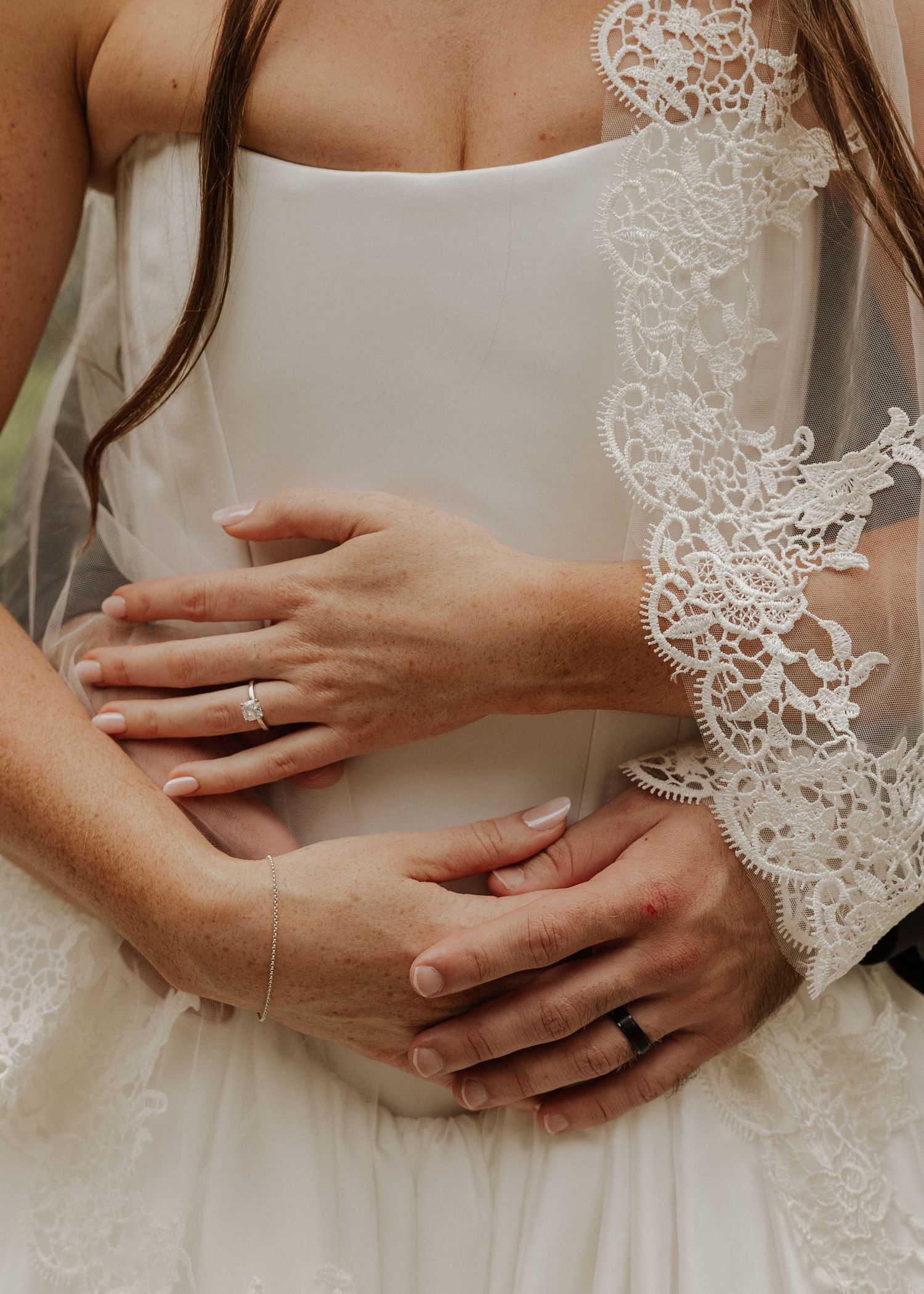 Close-up of bride and groom hands with wedding rings, lace veil detail visible on white bridal gown.