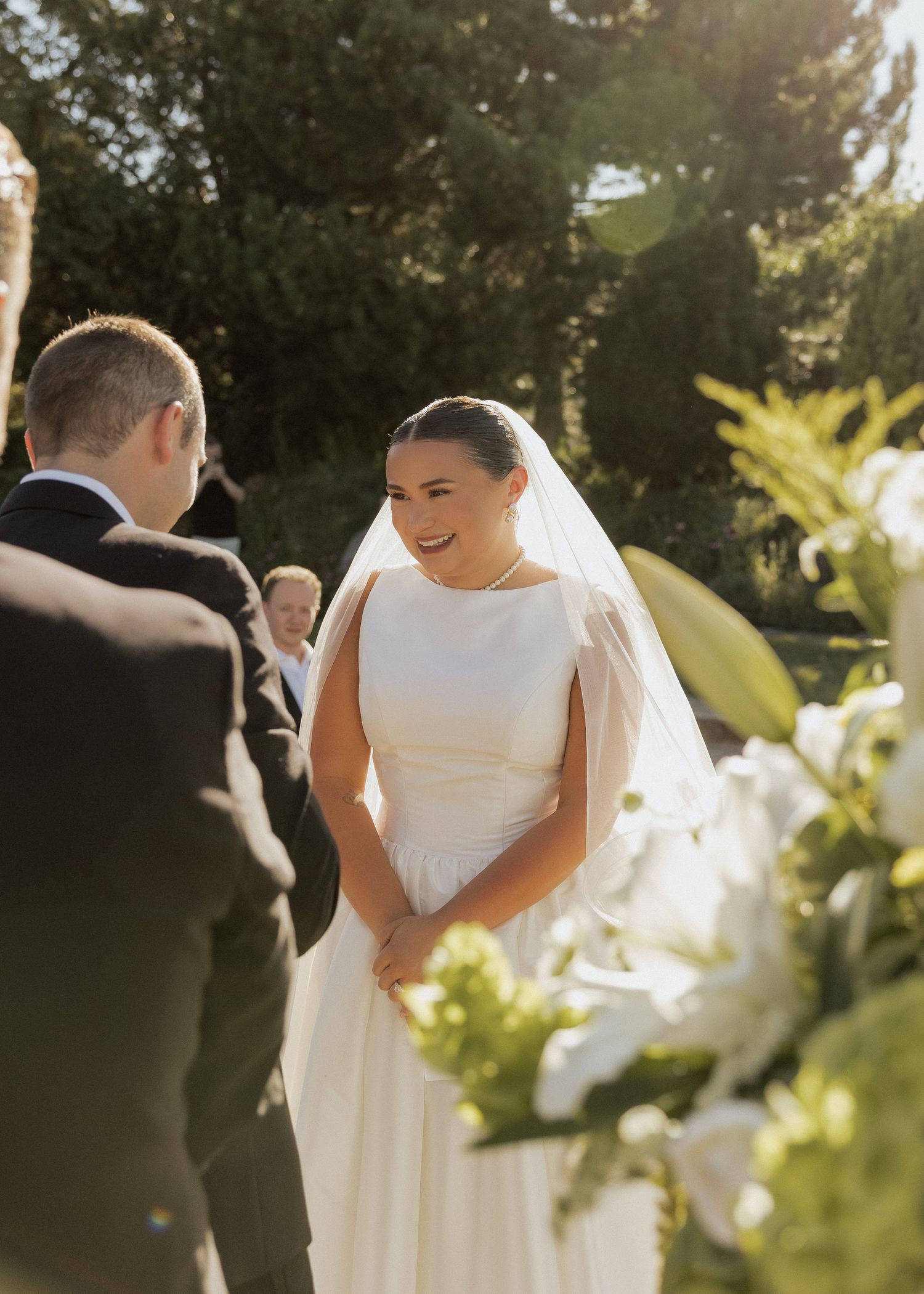 Bride in elegant white gown and veil smiles at groom during outdoor wedding ceremony.