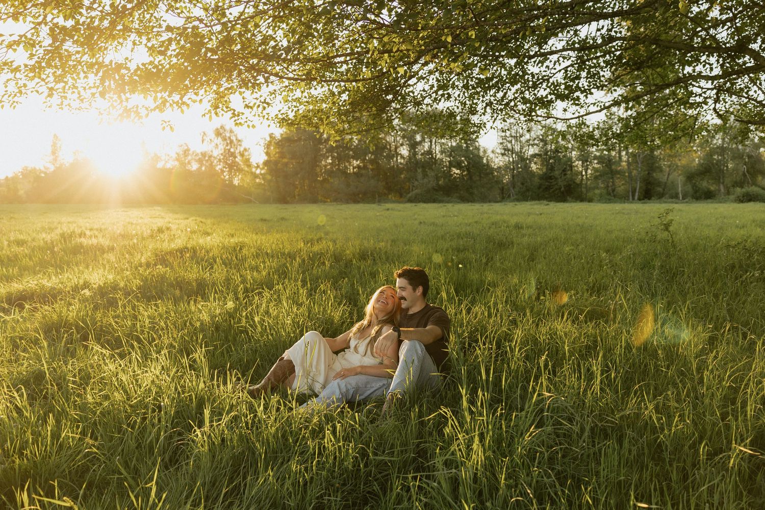 Couple sitting together in lush green meadow at golden sunset, embracing romantically under large tree in summer.