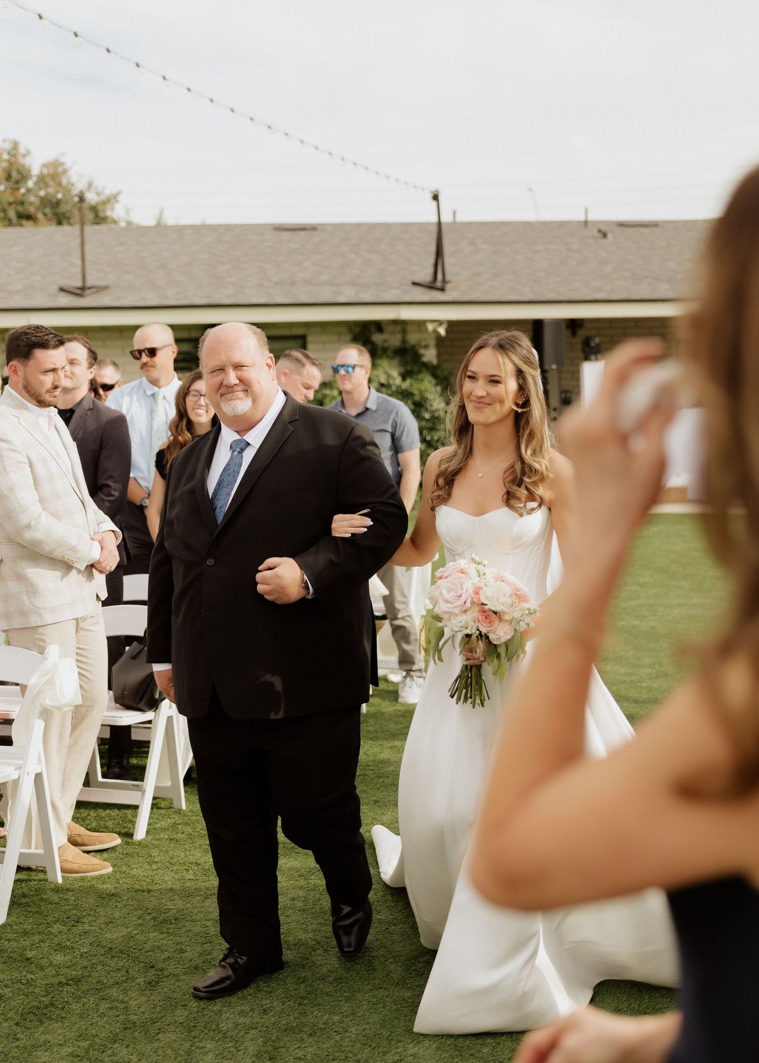 Bride in white gown walks down outdoor aisle with escort, holding pink bouquet, guests watching at sunny wedding ceremony.
