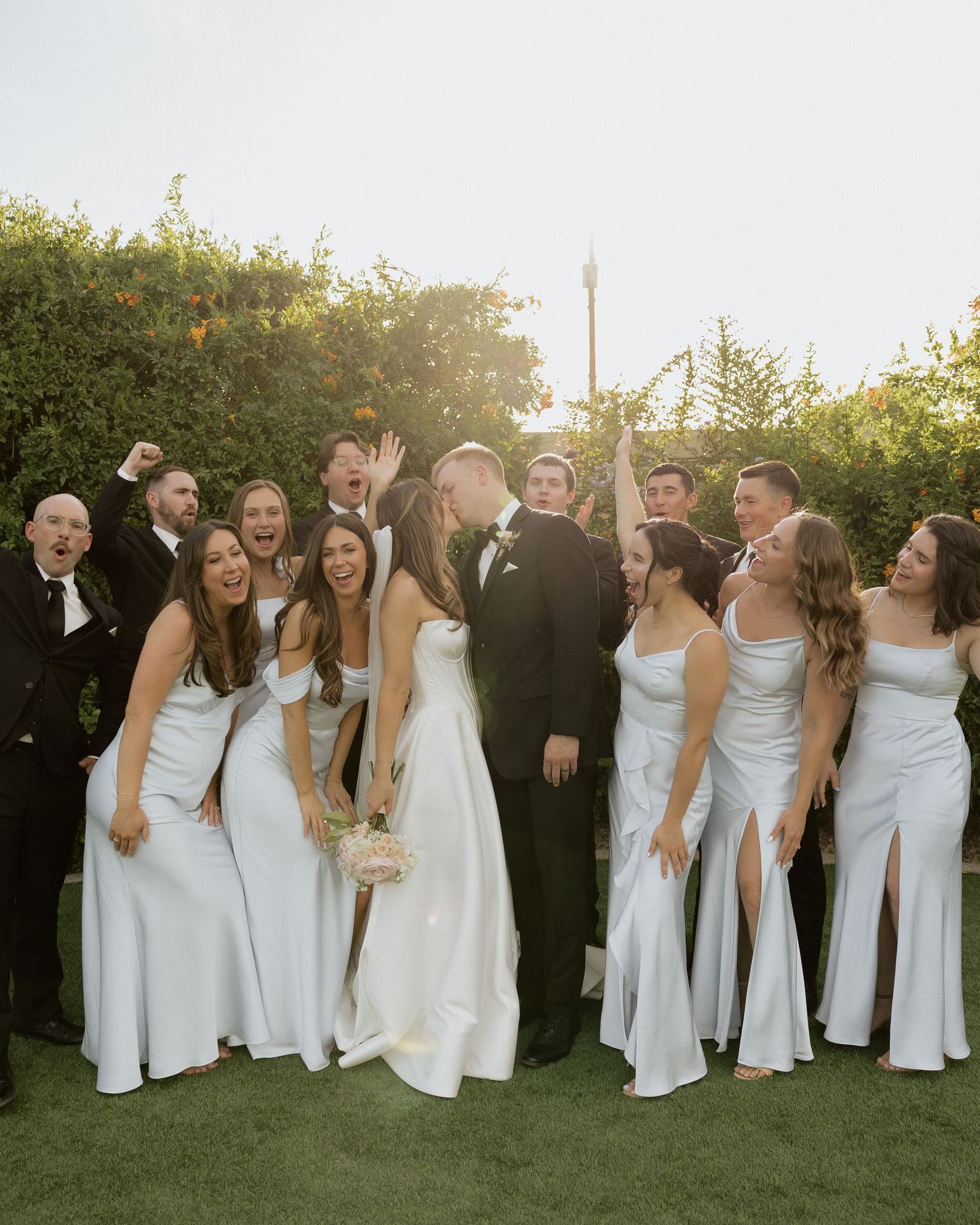 Wedding party celebrating outdoors, bride and groom kissing surrounded by bridesmaids in light blue gowns and groomsmen in tuxedos.