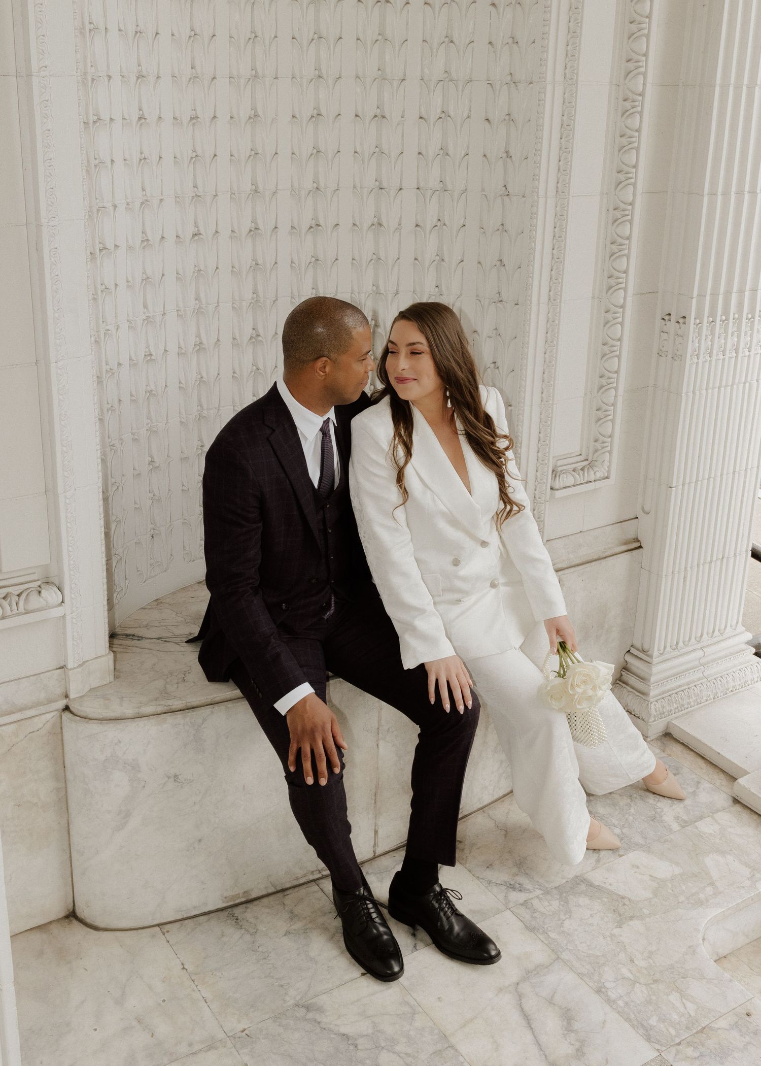 A couple in elegant black and white attire sits on marble steps, sharing a tender moment at a grand building.