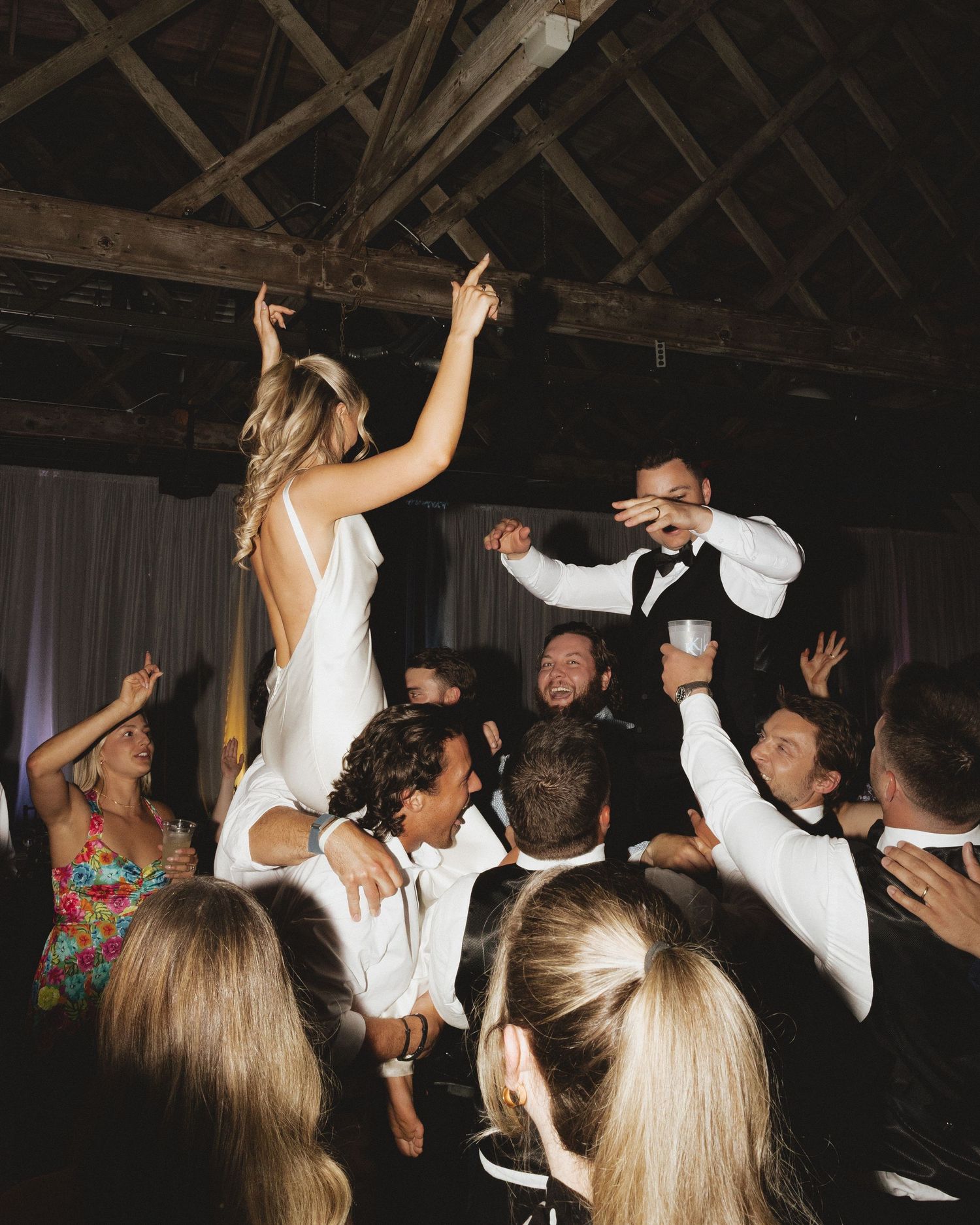 Bride in white dress raised by groomsmen on crowded wedding dance floor in rustic barn venue.