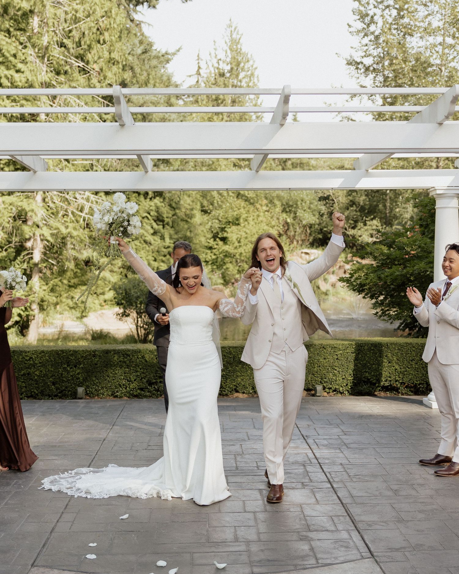 Joyful newlyweds celebrate recessional under white pergola surrounded by lush greenery.