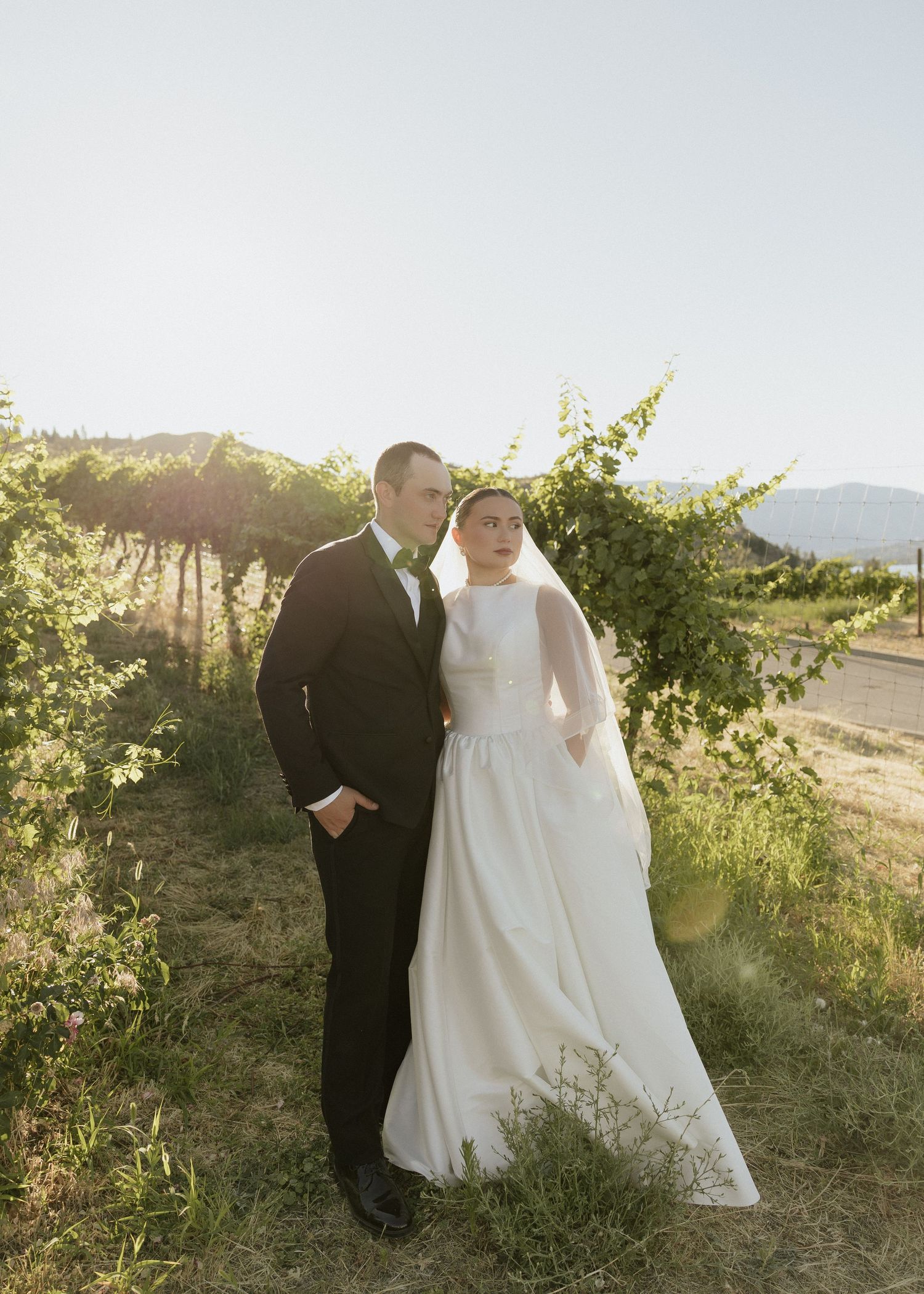 Bride and groom standing together in golden sunlight among lush vineyard rows at sunset.