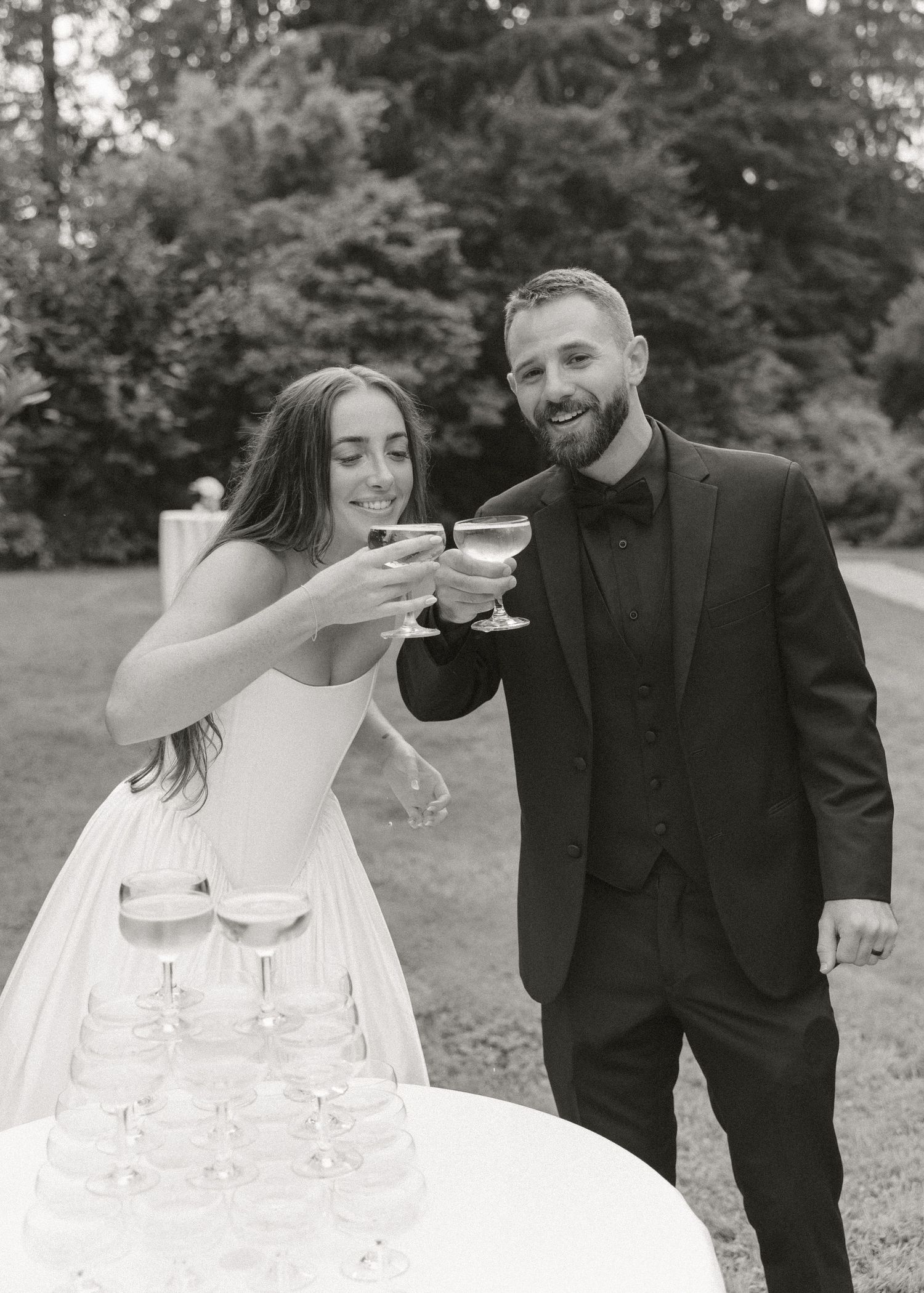 Bride and groom toasting champagne glasses at outdoor wedding reception beside a champagne tower, black and white photo.