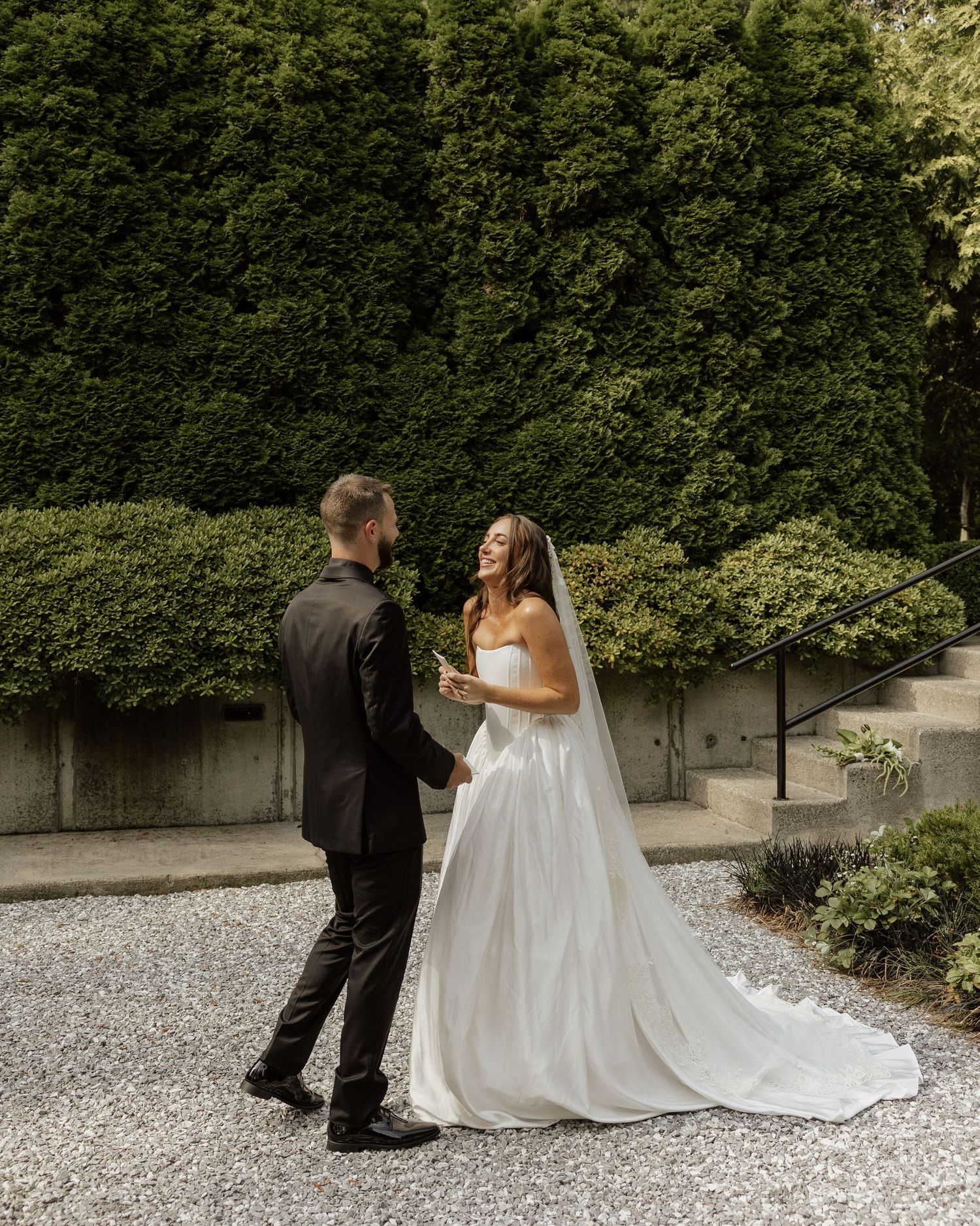 Bride in white strapless gown with veil shares first look moment with groom in black suit in garden.