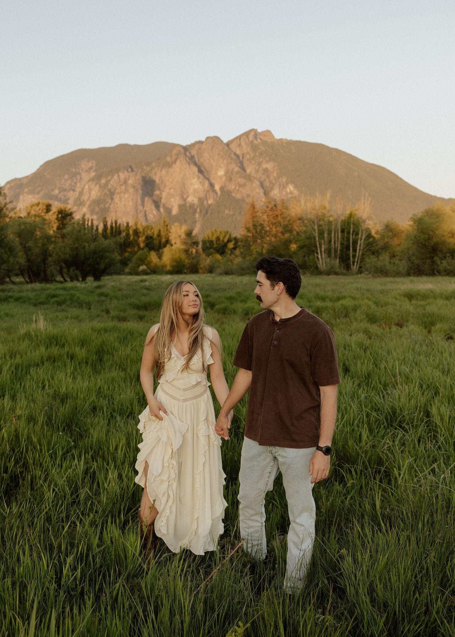 A couple holds hands in a green field with a dramatic mountain and golden sky behind them.