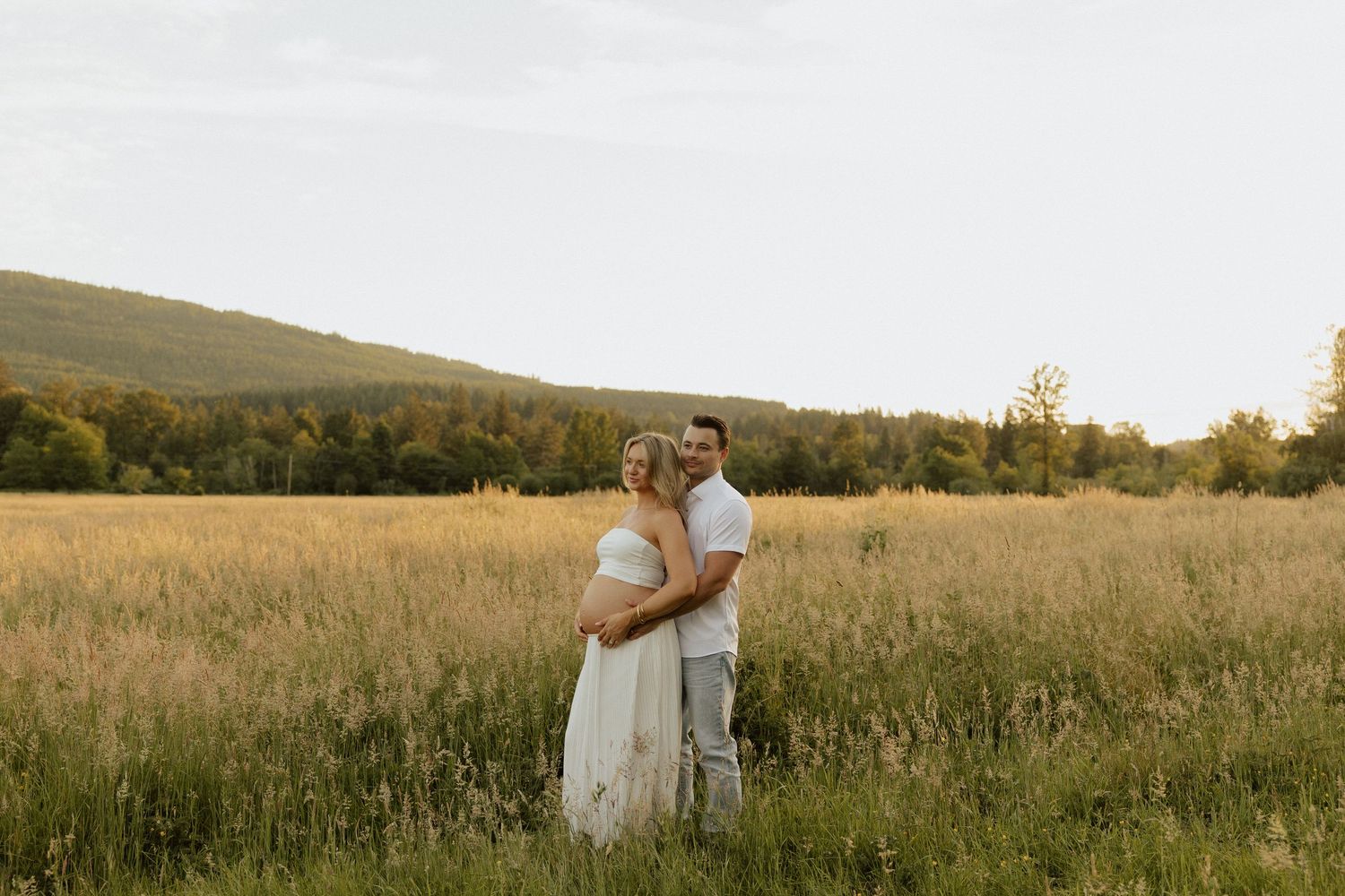A couple embraces in a golden field at sunset, with lush green hills and trees in the background.