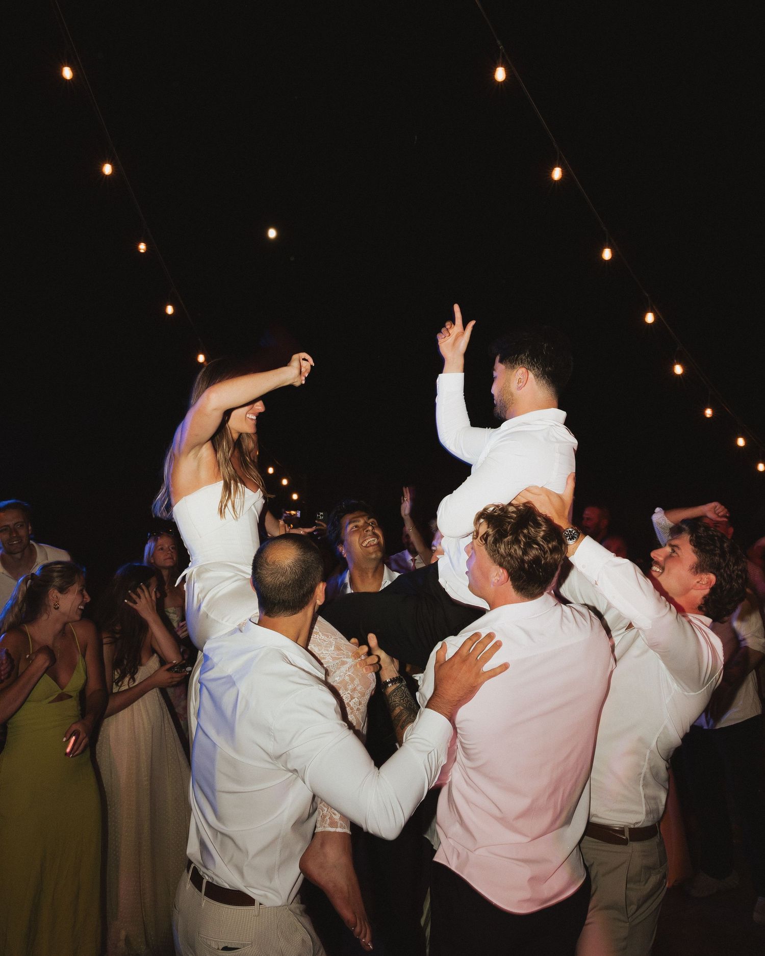 Bride and groom lifted by guests during lively outdoor wedding reception dance under string lights.