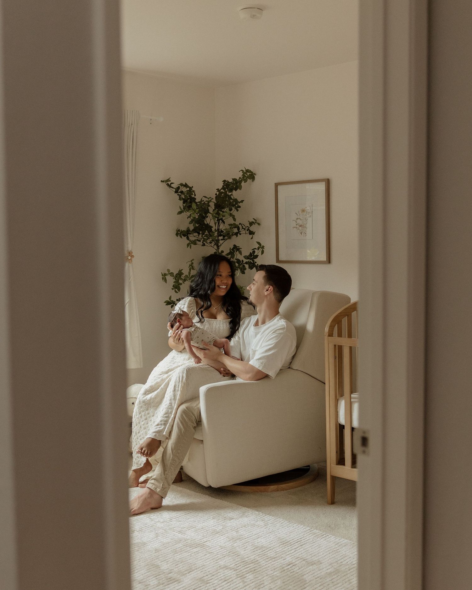 Parents sharing tender moment with newborn in white nursery room, viewed through doorframe.