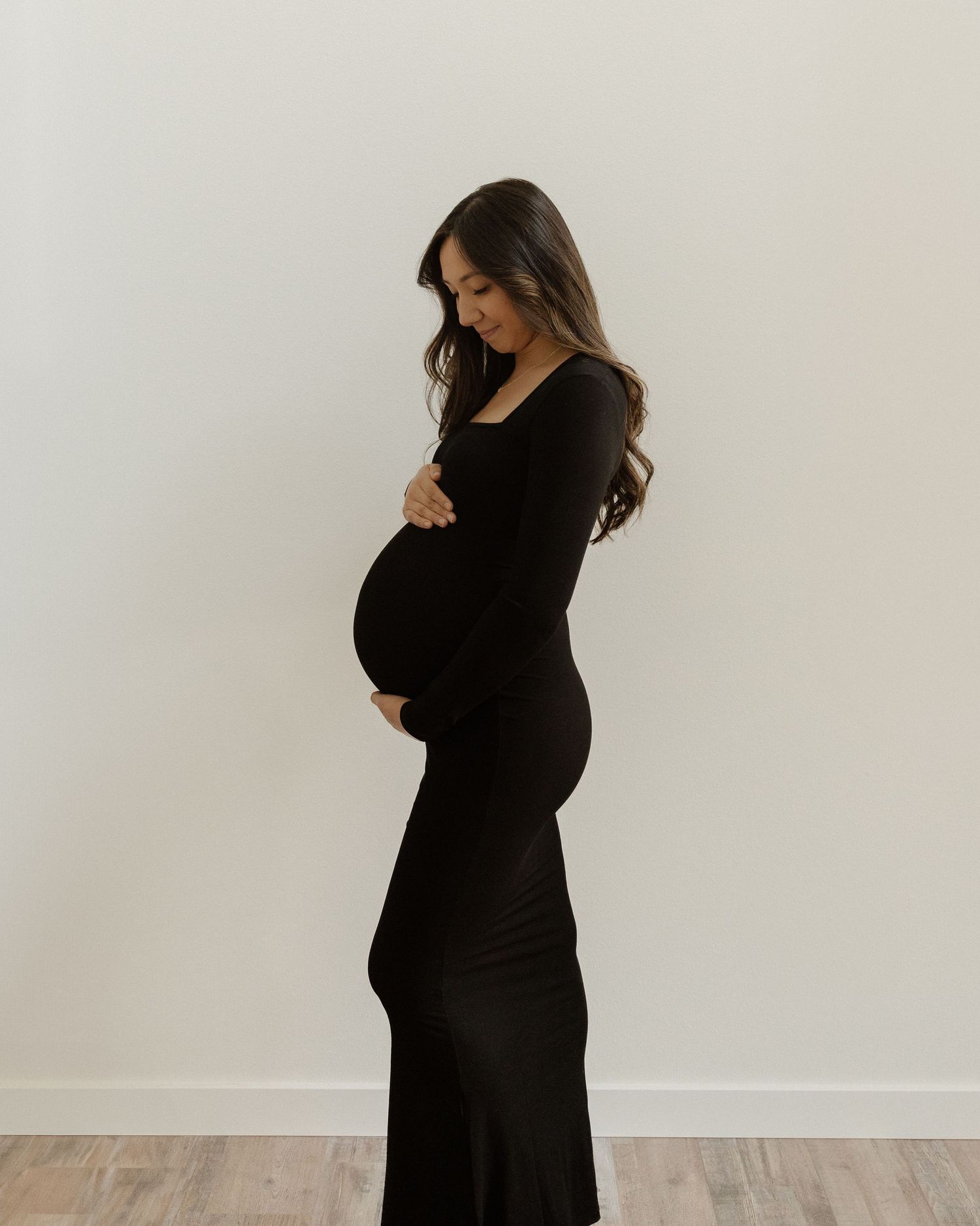 Expectant mother in a fitted black long dress poses elegantly against a white wall.
