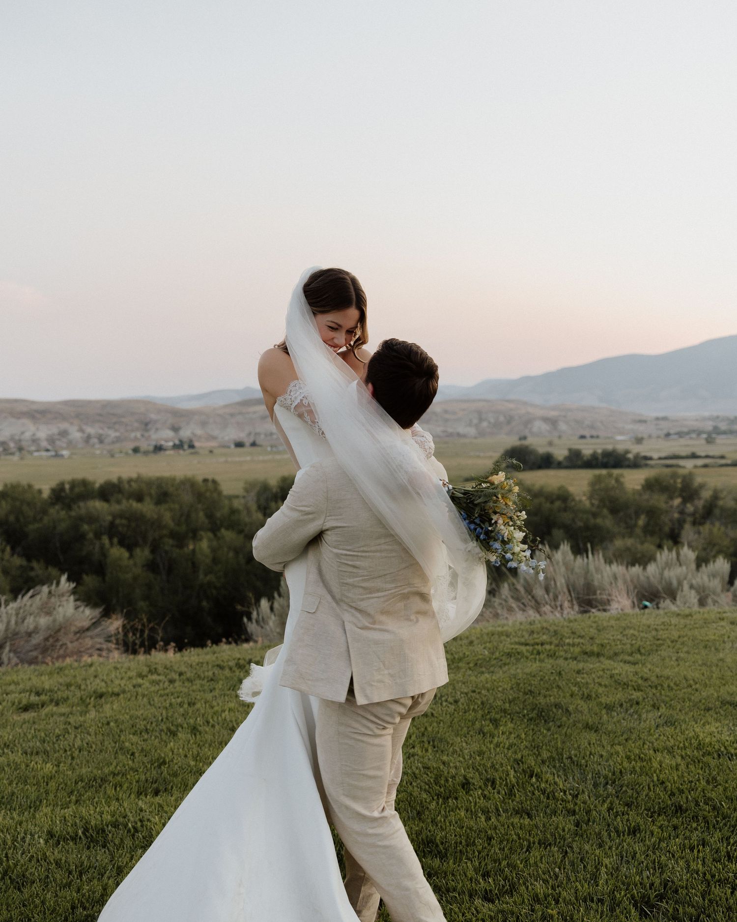 Groom lifts bride in romantic embrace on grassy hilltop with mountain landscape in background.