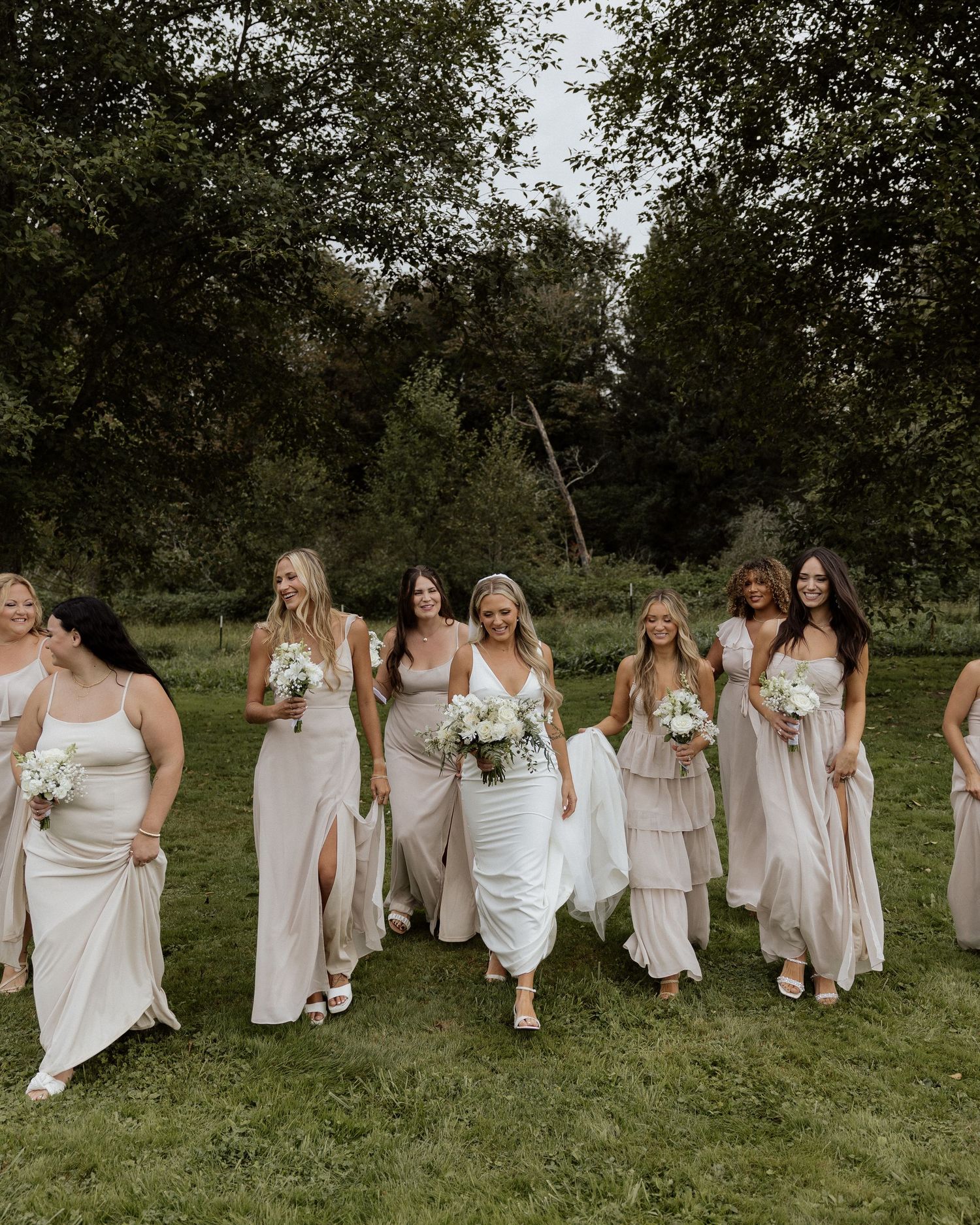 Bridal party in neutral-toned dresses walks together across a green field carrying white floral bouquets.
