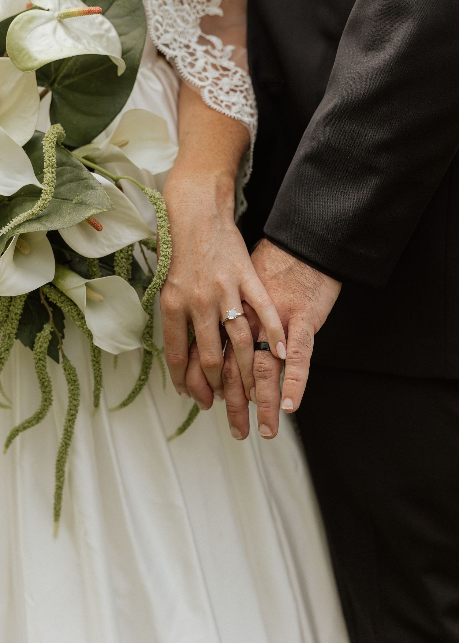 Close-up of bride and groom holding hands, showing wedding rings, with white calla lily bouquet.