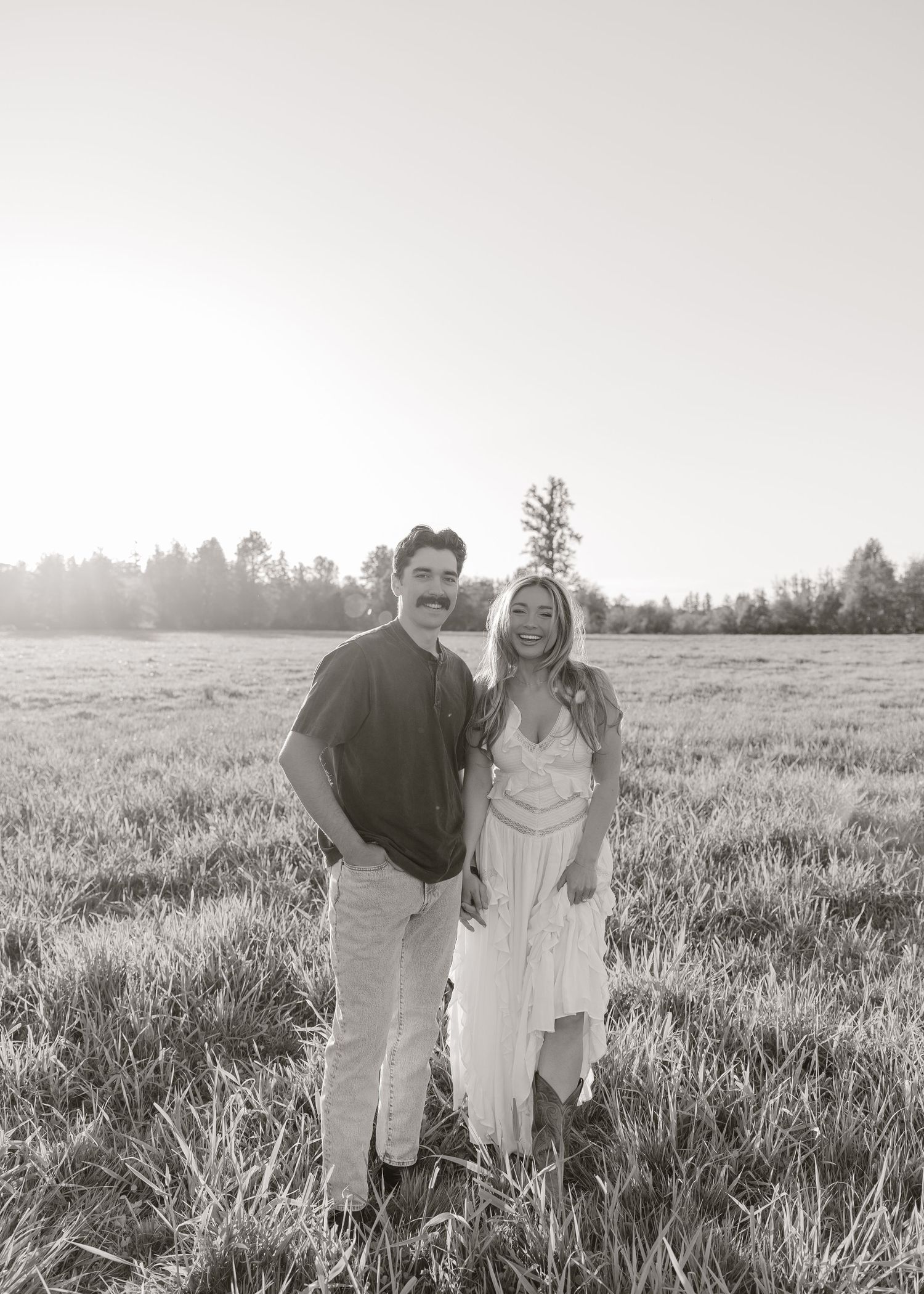 Black and white photo of a smiling couple standing in an open field with trees in the background.