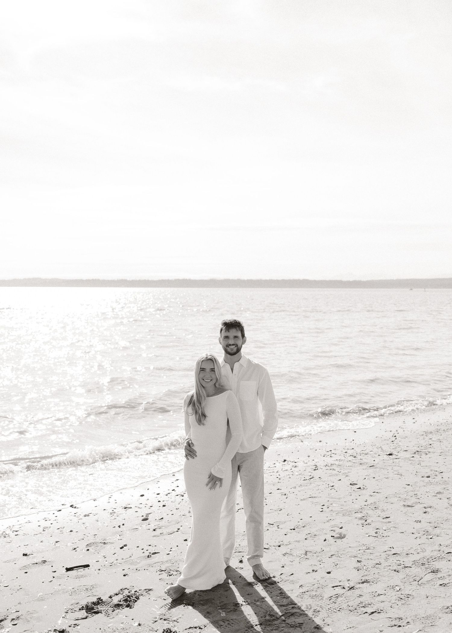 Black and white beach photo of a smiling couple in white outfits standing by the ocean shoreline.