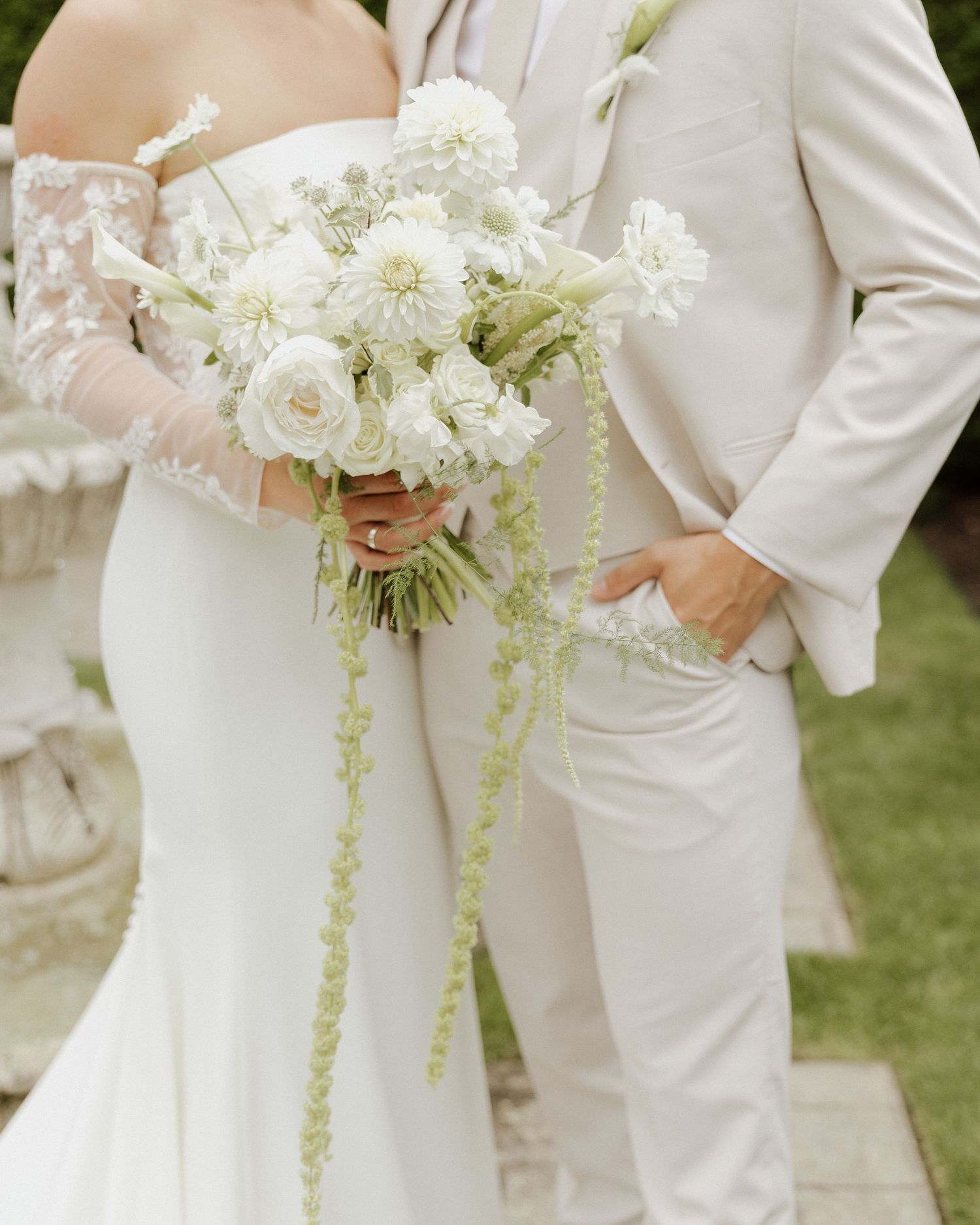 Bride holds a cascading white bouquet with greenery beside groom in a beige suit at an outdoor wedding.