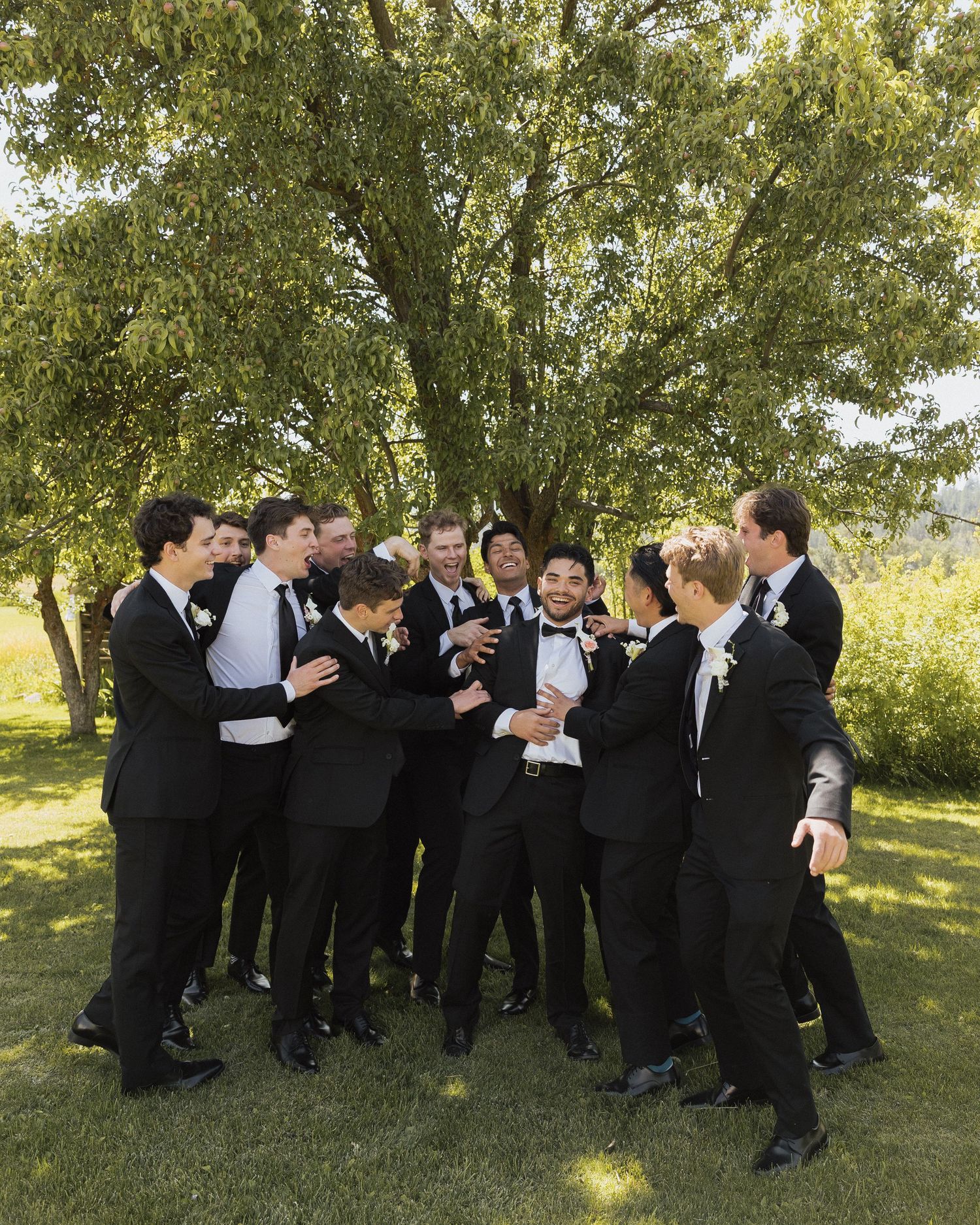 Groom surrounded by groomsmen in black tuxedos laughing outdoors under large tree at wedding.