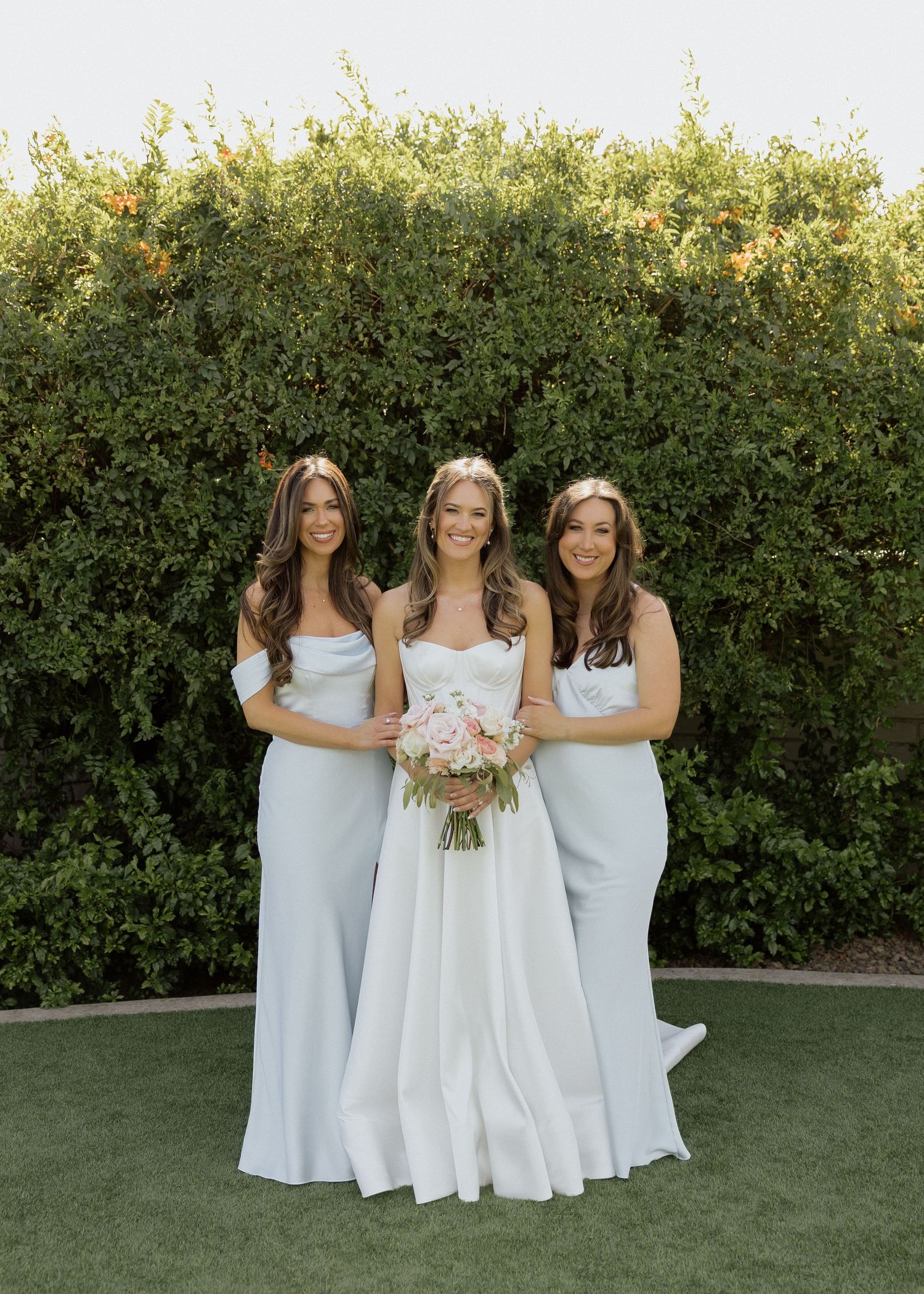 Bride in white strapless gown holding pink bouquet flanked by two bridesmaids in light blue dresses outdoors.