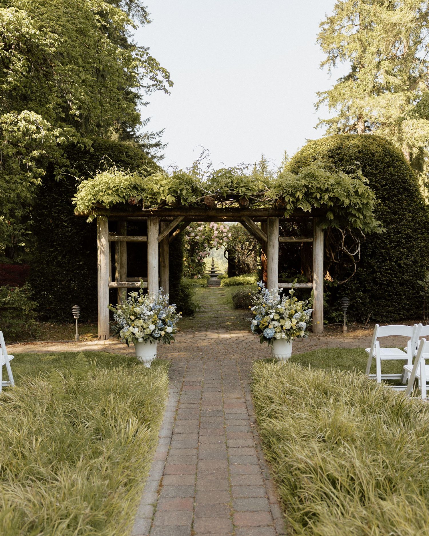 Elegant outdoor wedding ceremony aisle with brick path, floral arrangements, and vine-covered wooden pergola surrounded by lush greenery.