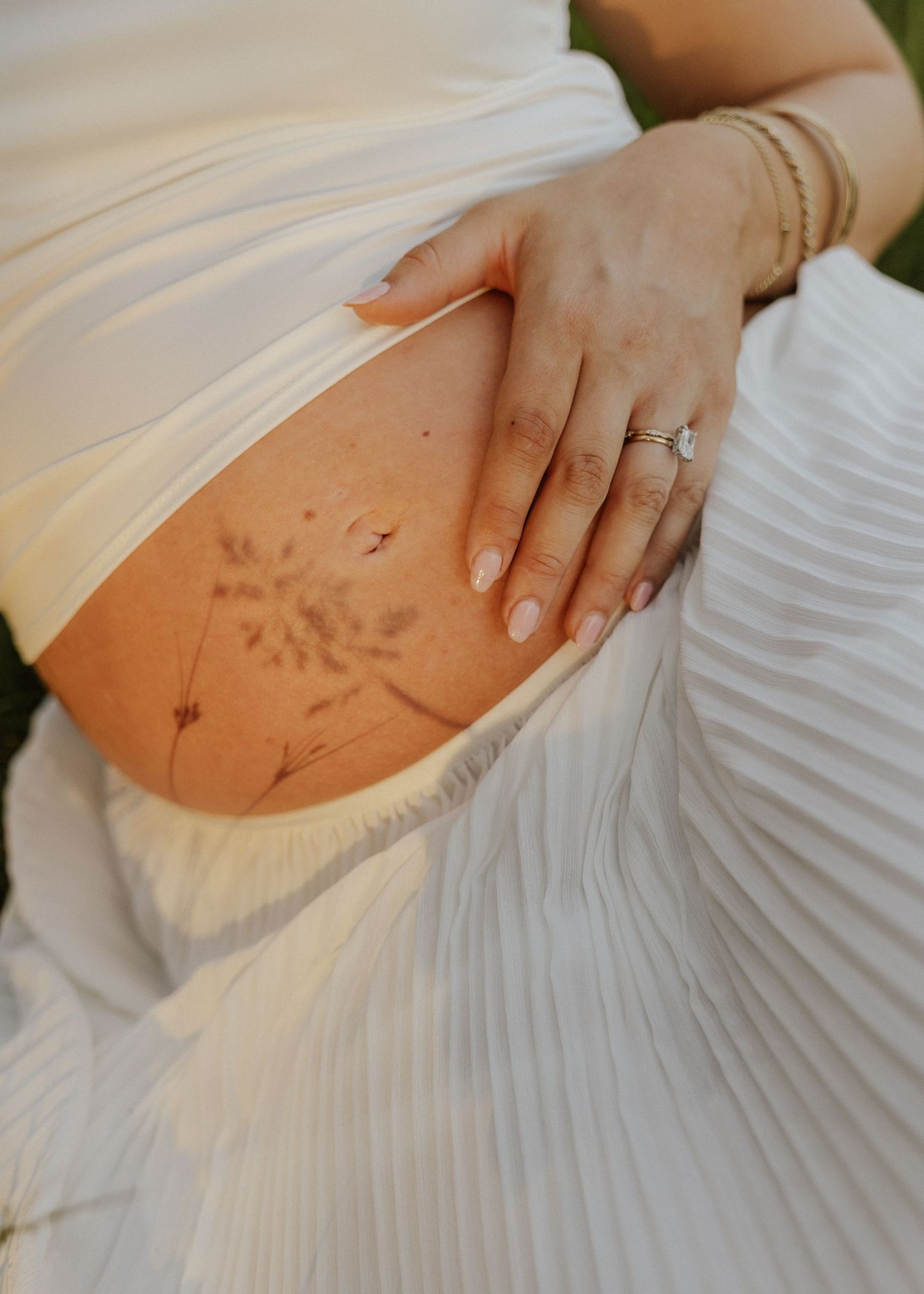 Close-up of pregnant woman's baby bump in white pleated skirt, wearing a diamond ring and gold bracelets.