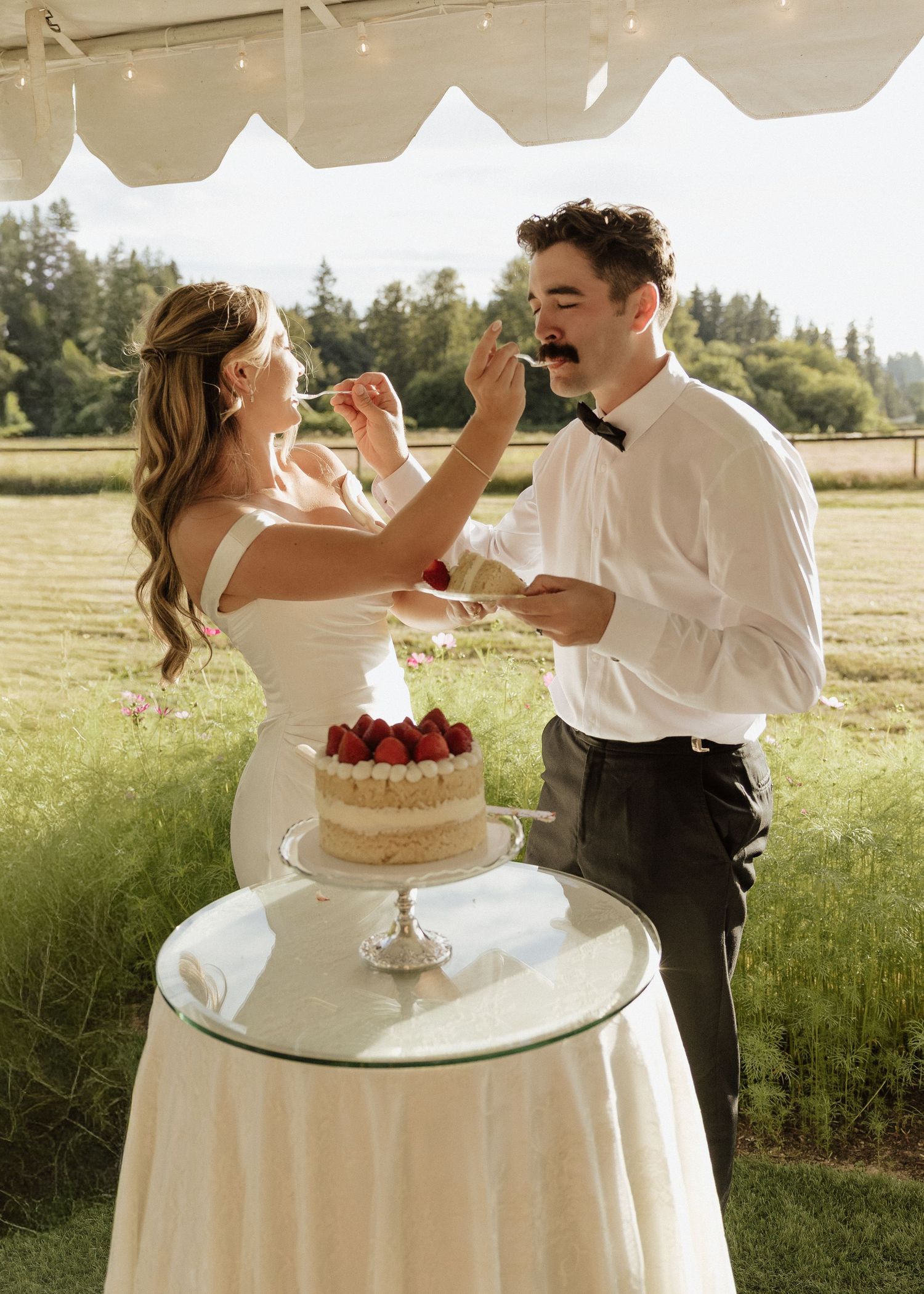 Bride and groom playfully feed each other cake at outdoor wedding with strawberry-topped white naked cake.