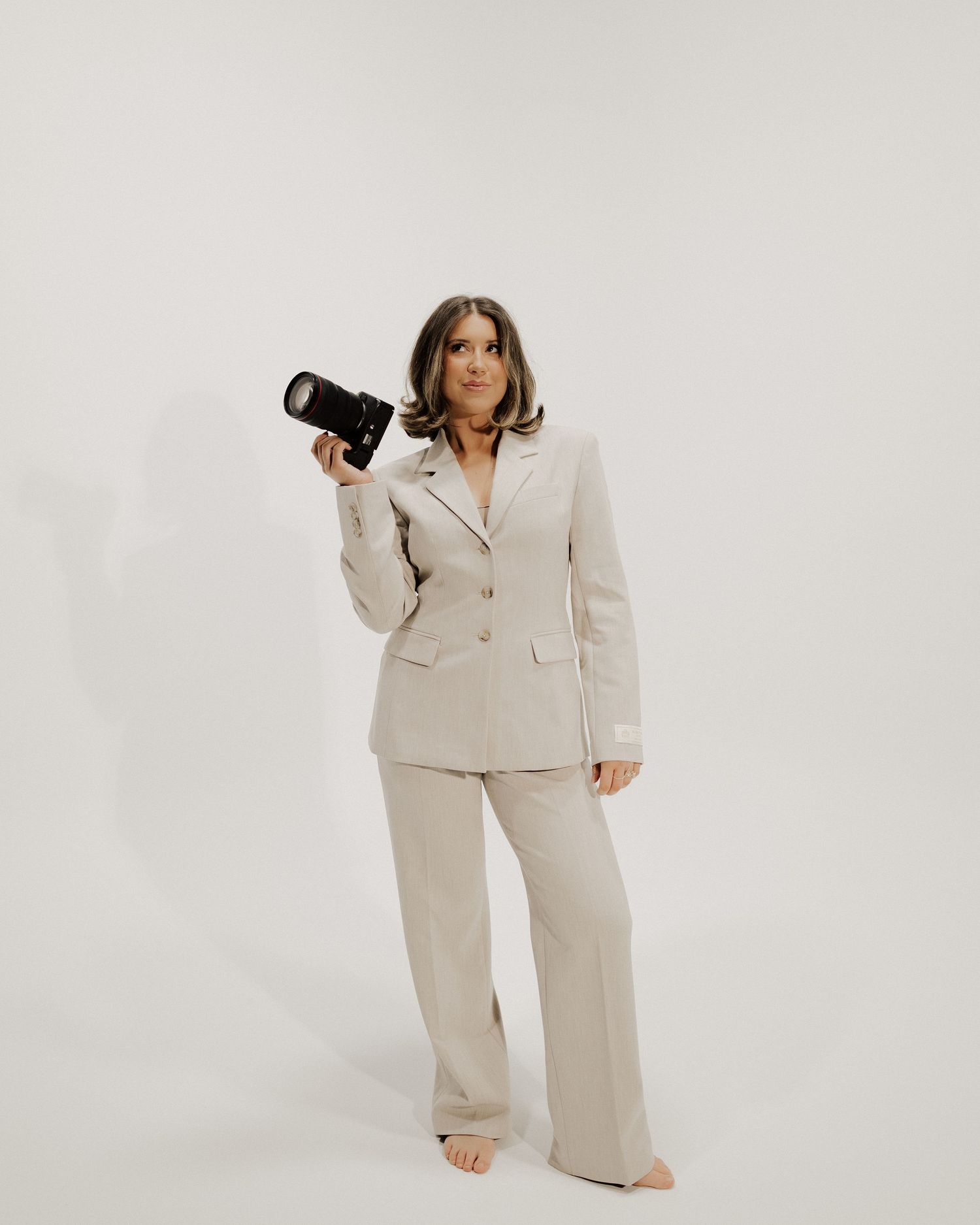 Female photographer in stylish beige suit holding professional camera in studio setting.