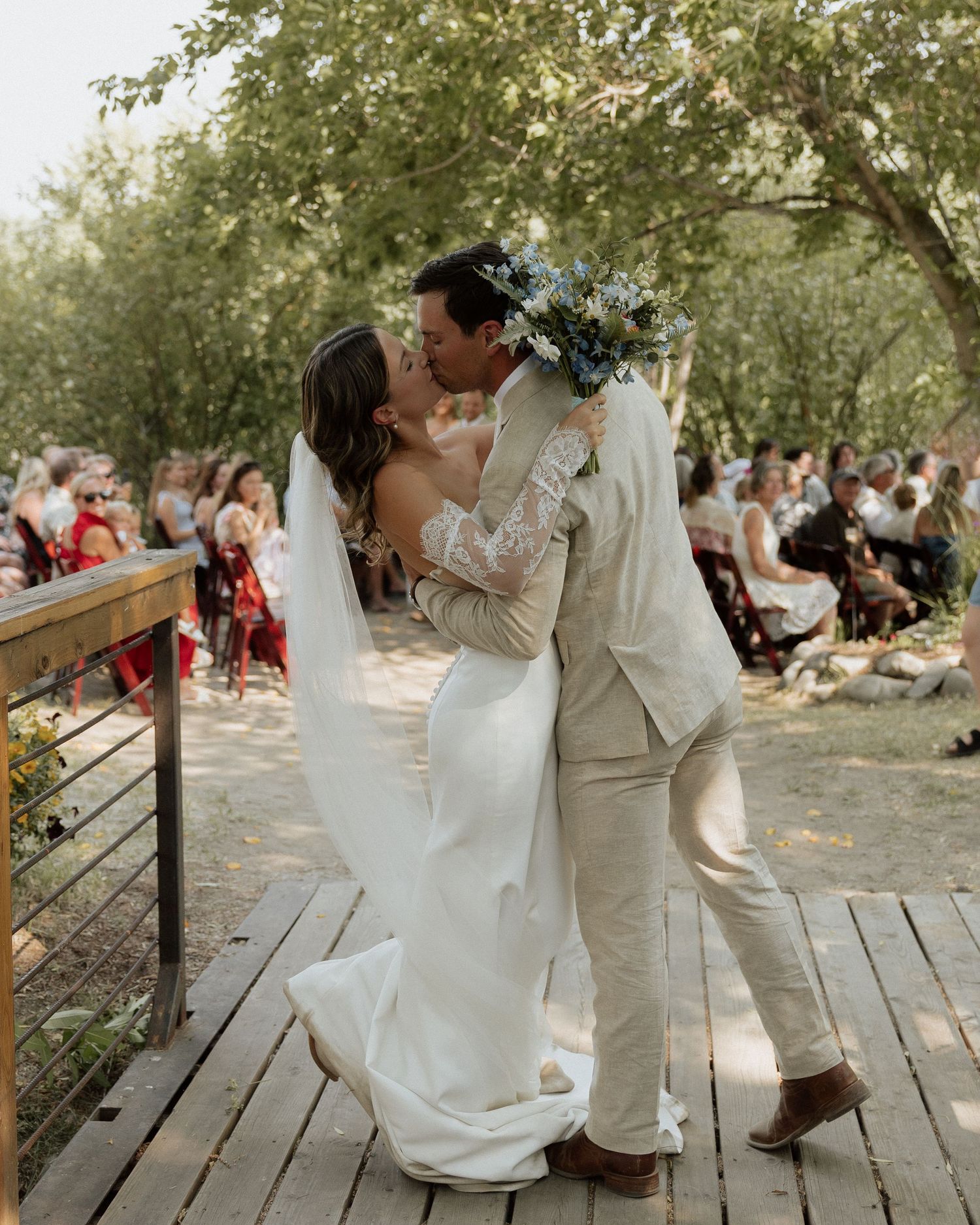 A passionate kiss between newlyweds on a wooden deck surrounded by wedding guests.