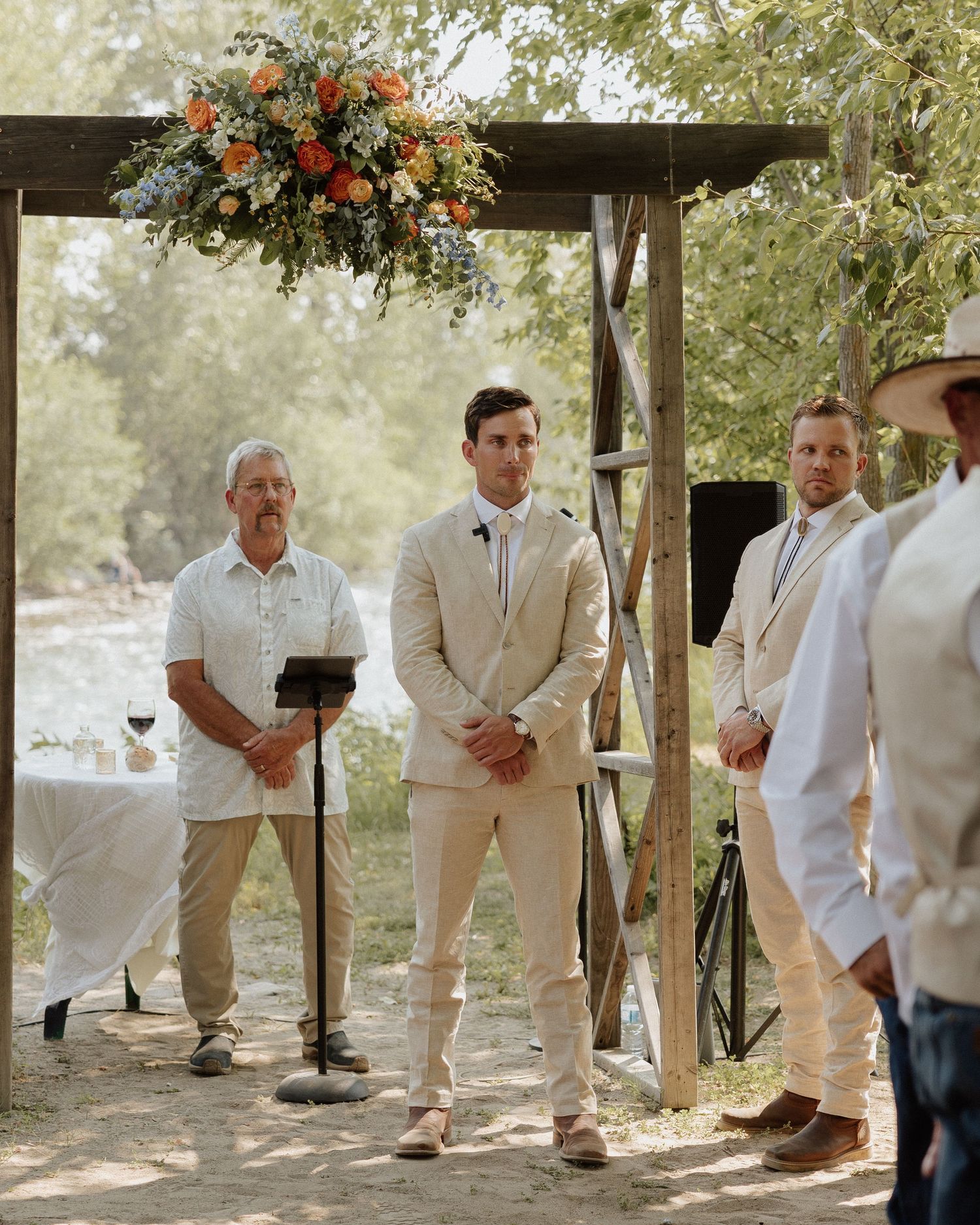 Wedding ceremony setup with floral arch and wedding party in light colored suits standing at altar.
