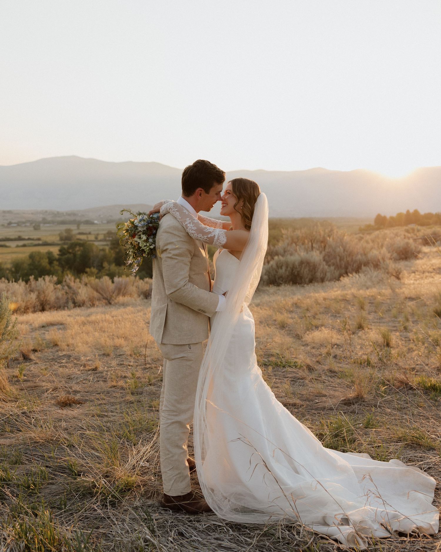 Romantic sunset portrait of newlyweds embracing on hilltop with mountain landscape.