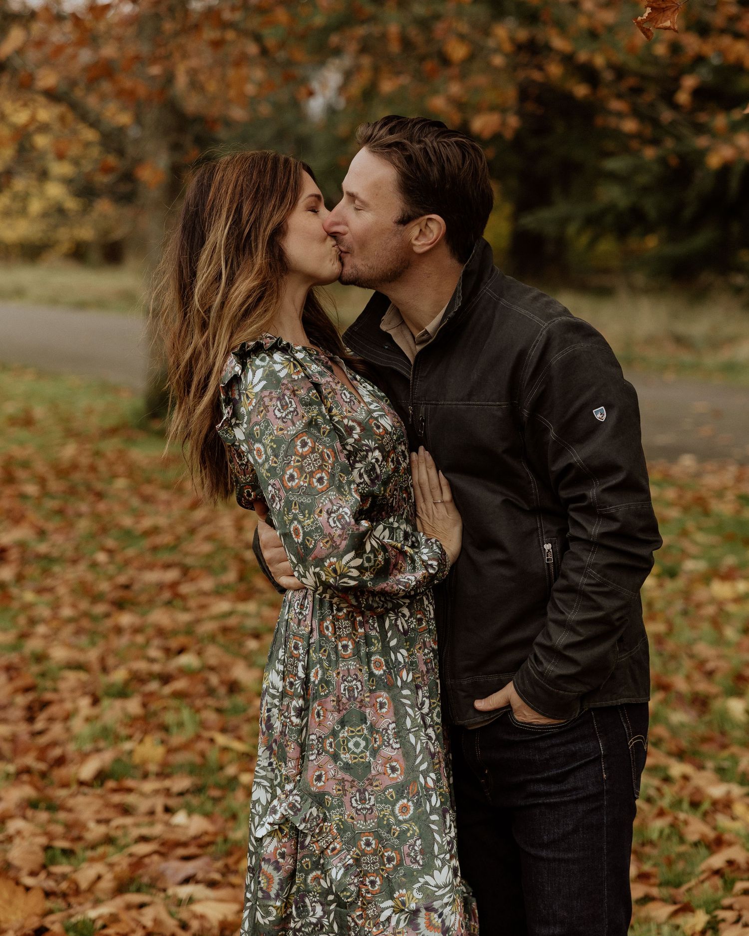 A romantic couple shares a kiss in a park surrounded by fall foliage.