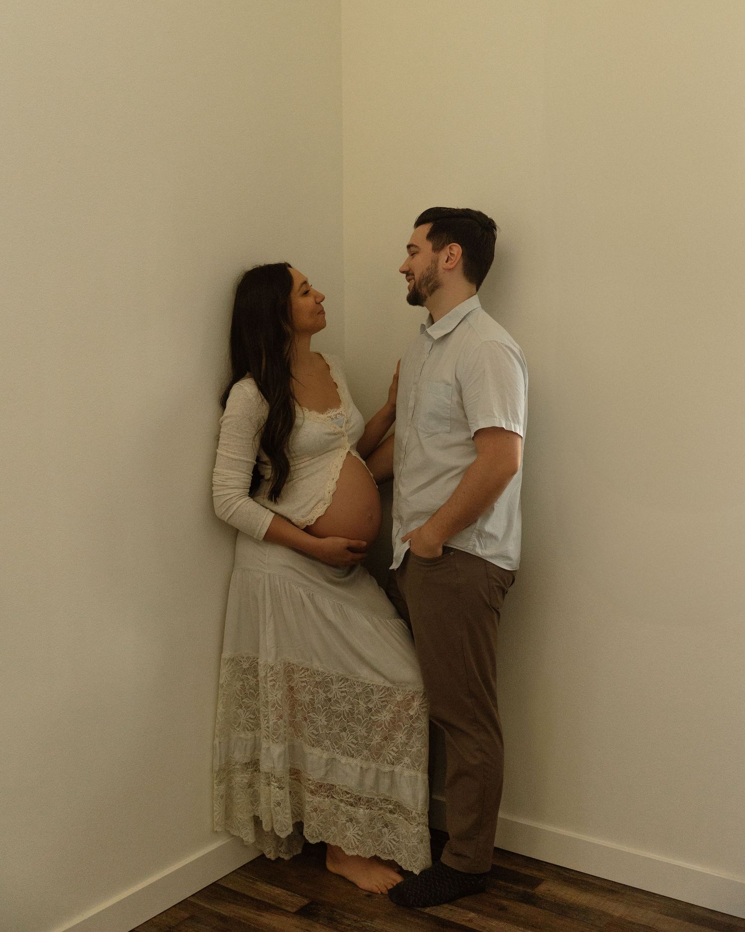 An intimate maternity photoshoot of a couple standing against a minimalist beige wall.