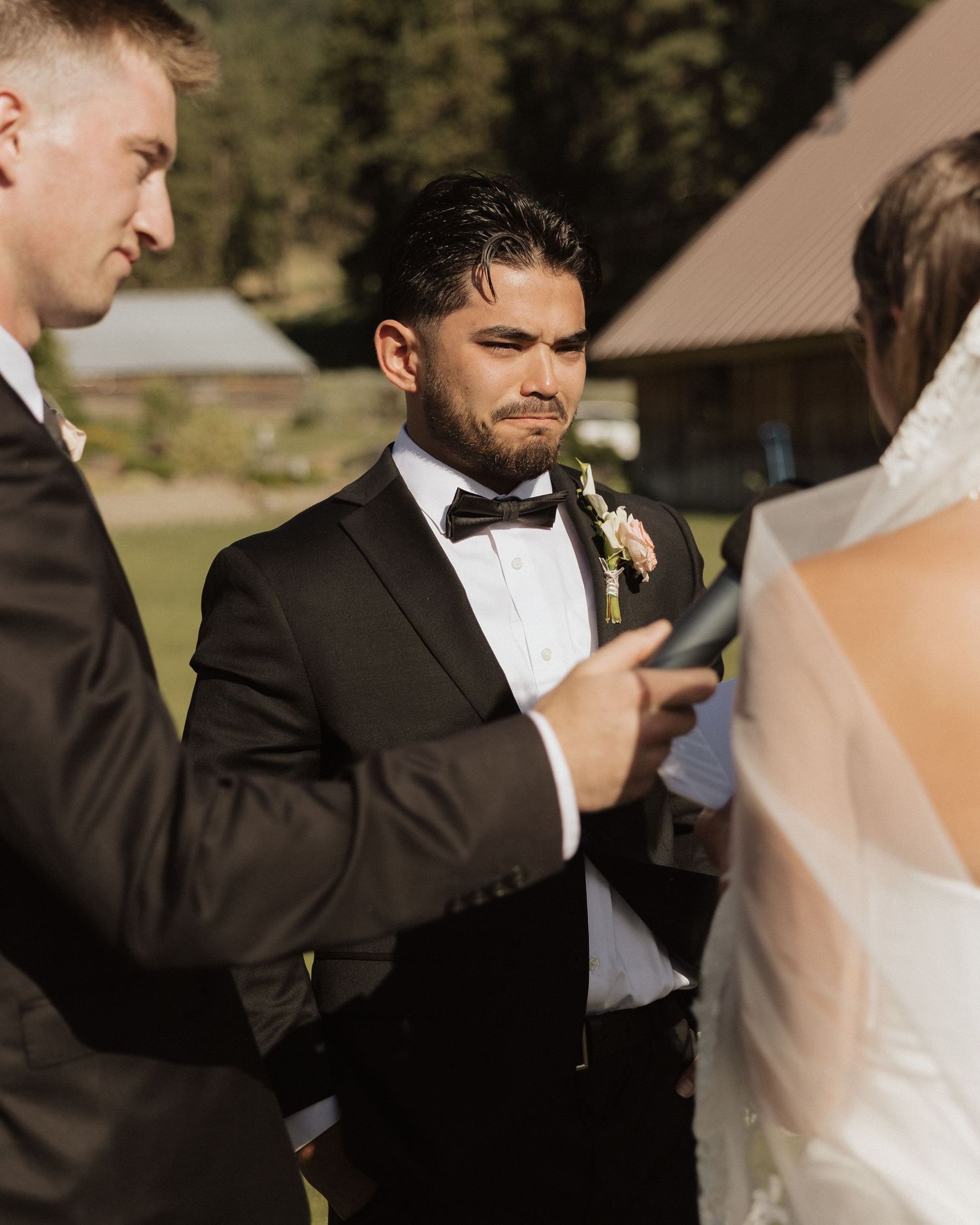 Emotional groom in black tuxedo with bow tie listens to vows during outdoor wedding ceremony.