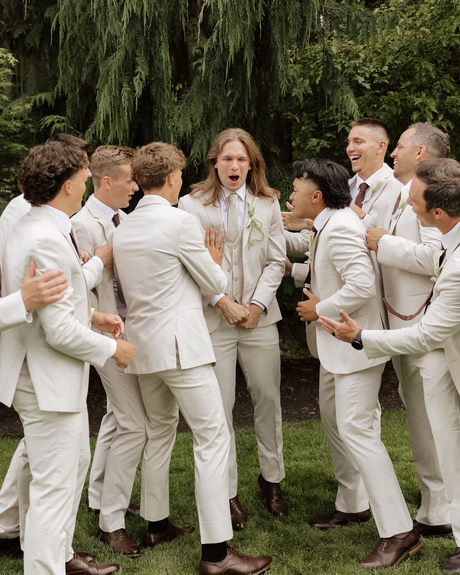 Groomsmen in cream suits surround and celebrate the groom outdoors at a wedding.