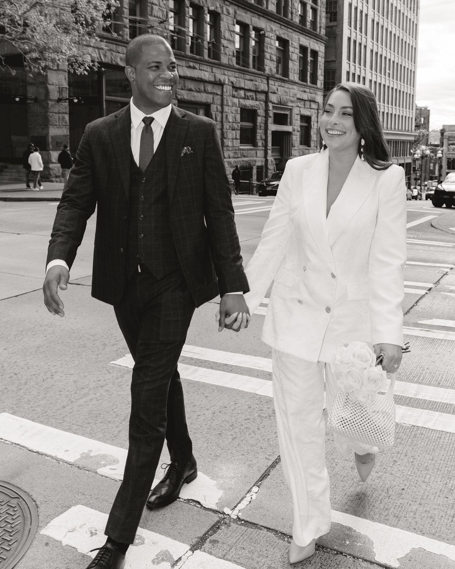 Elegant couple in black and white photo crossing a city street, man in dark suit, woman in white blazer suit.