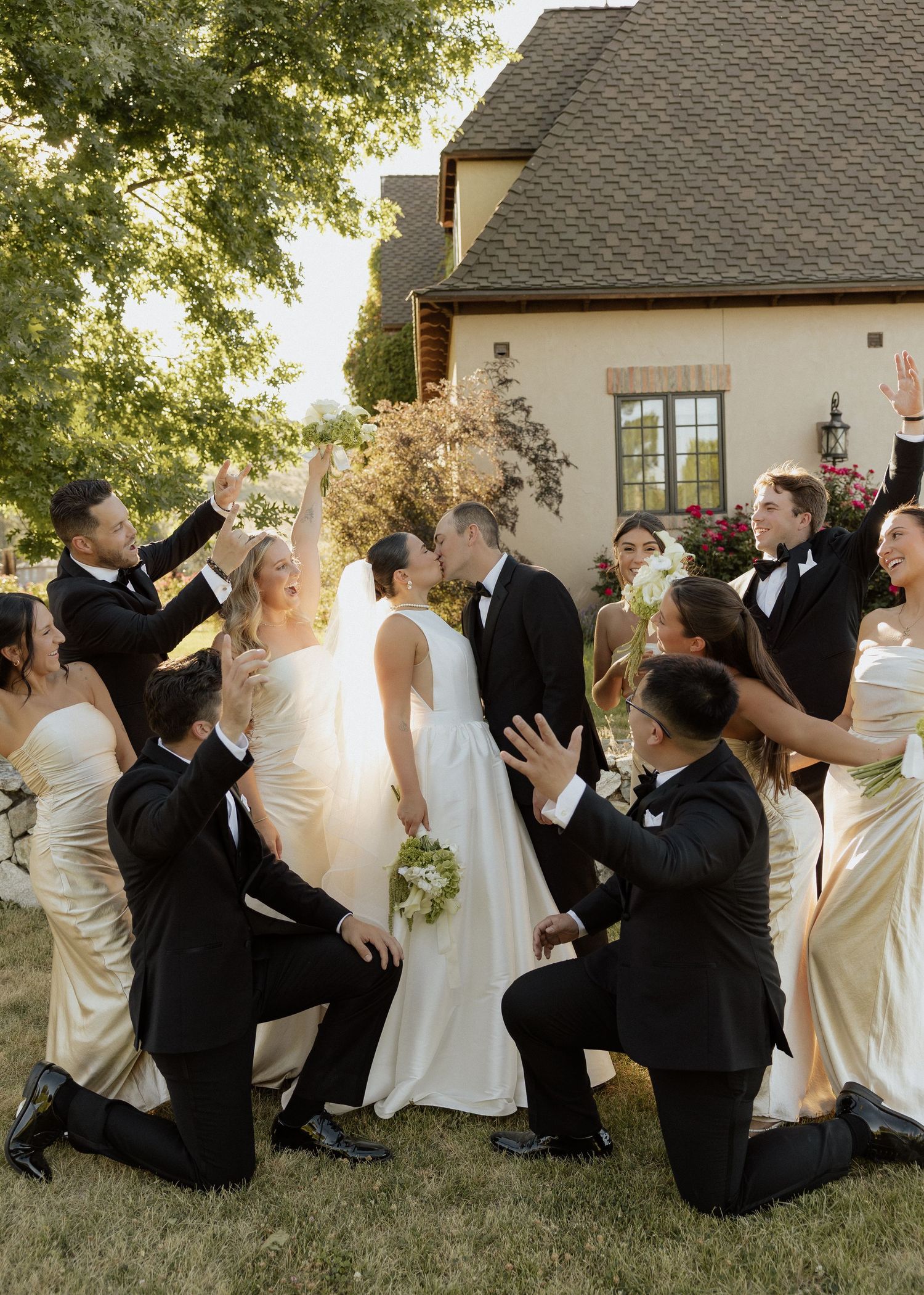 Wedding party celebrating bride and groom outdoors with cheering and kneeling near elegant venue.