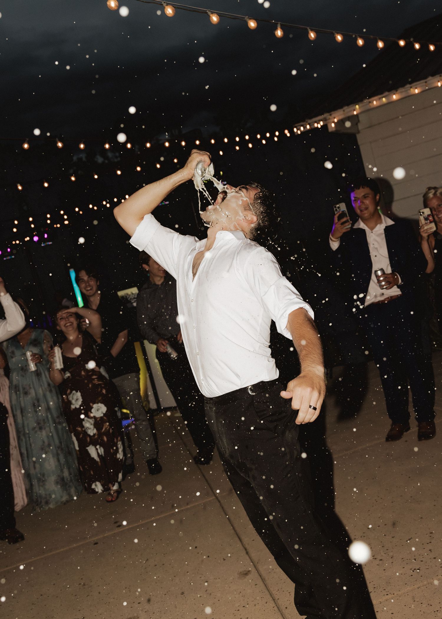 Groom in white shirt pouring champagne into his mouth at outdoor wedding reception with string lights.