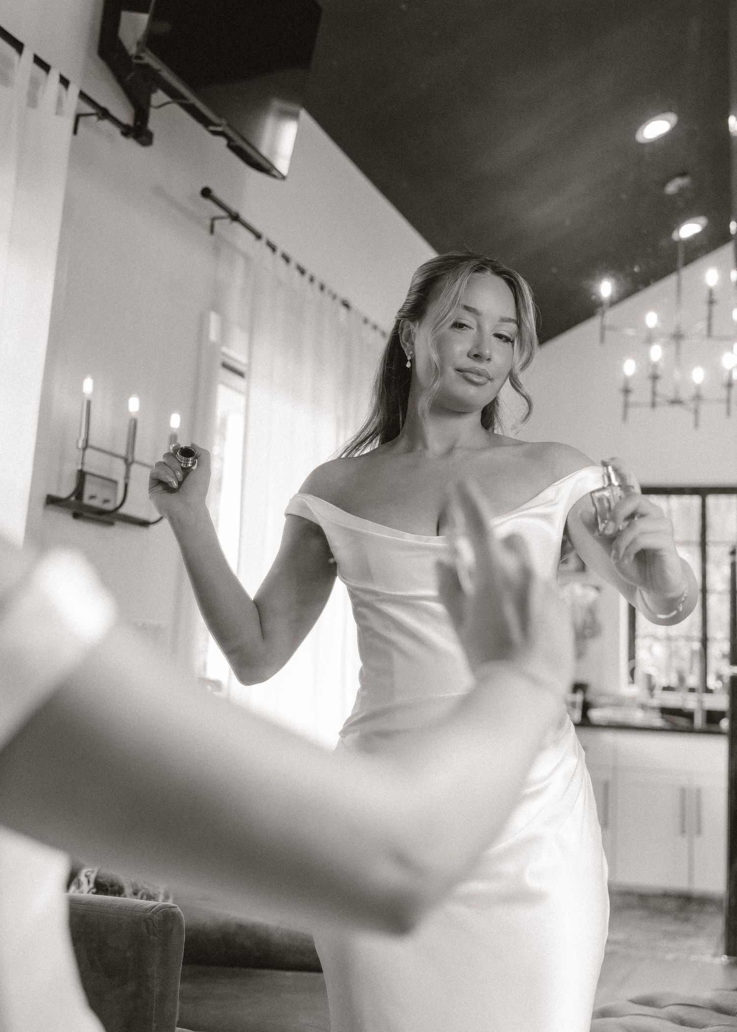 Black and white photo of a bride in an off-shoulder white gown holding a perfume bottle, smiling in an elegant room.