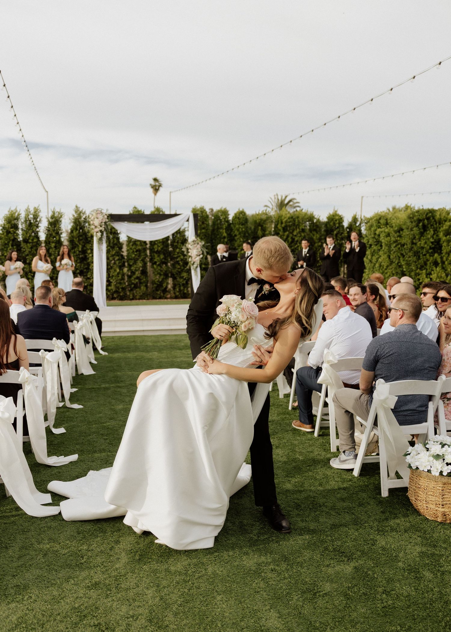 Bride and groom share a kiss during outdoor wedding ceremony on green lawn with white chairs and floral arch backdrop.