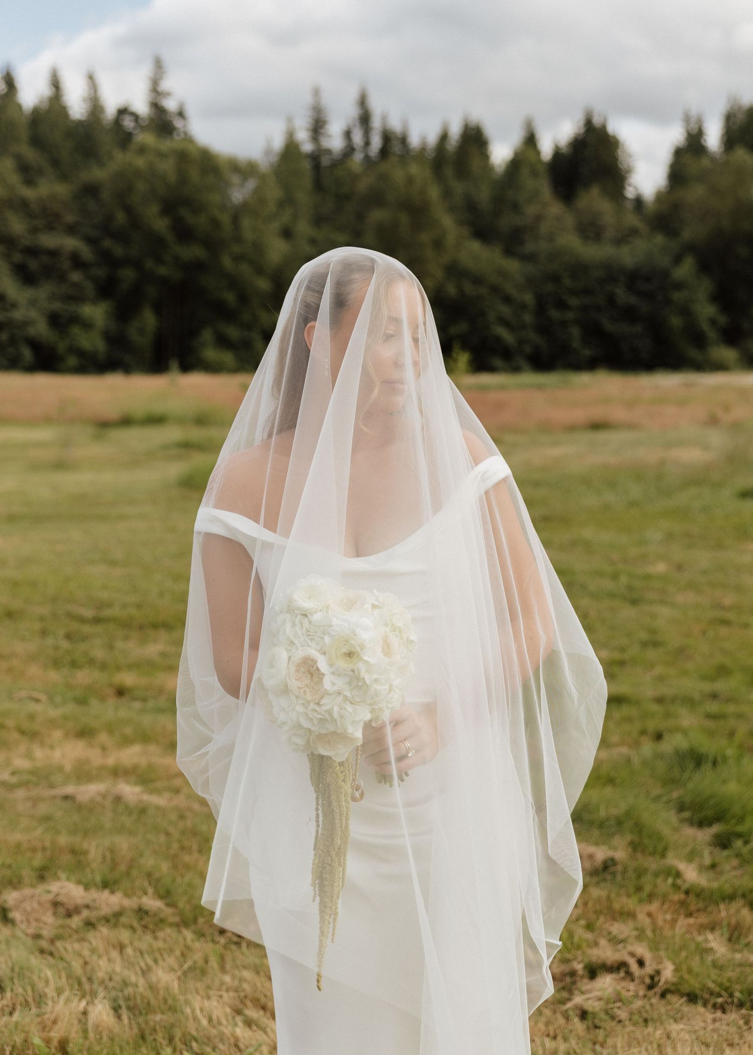Bride in white gown and flowing veil holding bouquet of white flowers in a green field.
