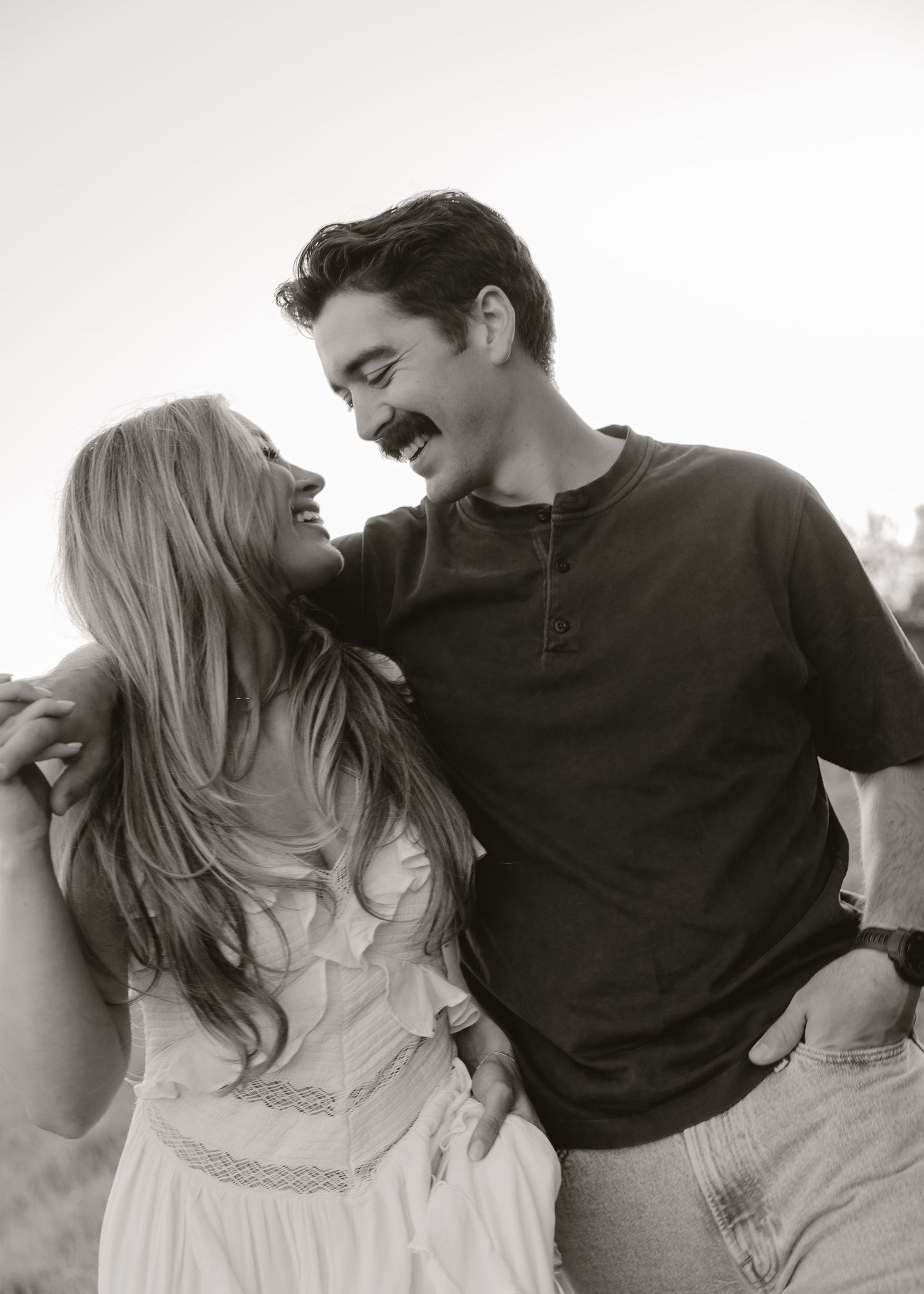 Black and white photo of a laughing couple embracing outdoors, woman in white dress, man in dark henley shirt.