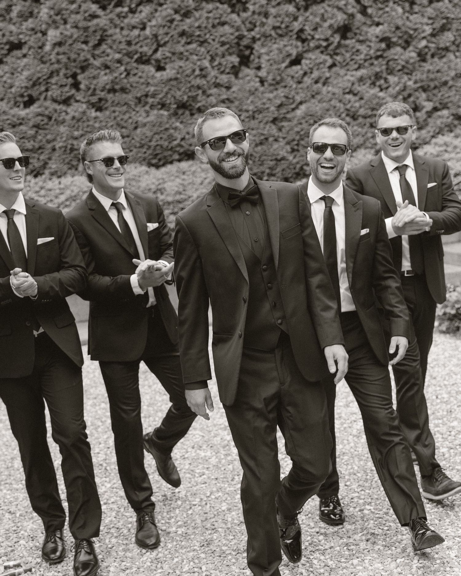 Black and white photo of a groom and groomsmen in dark suits and sunglasses walking and laughing outdoors.
