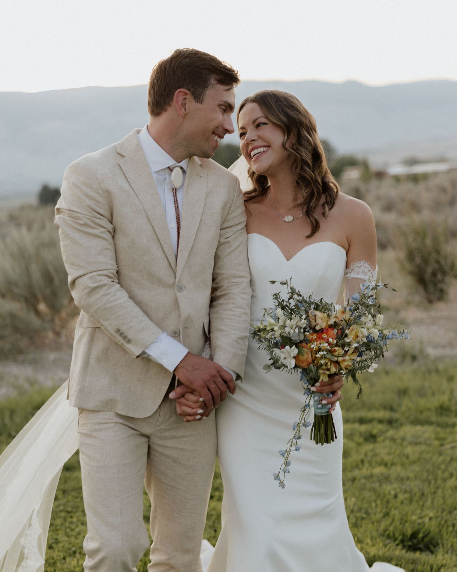 Newlyweds share intimate moment outdoors with bride holding rustic bouquet in strapless gown and groom in beige suit.