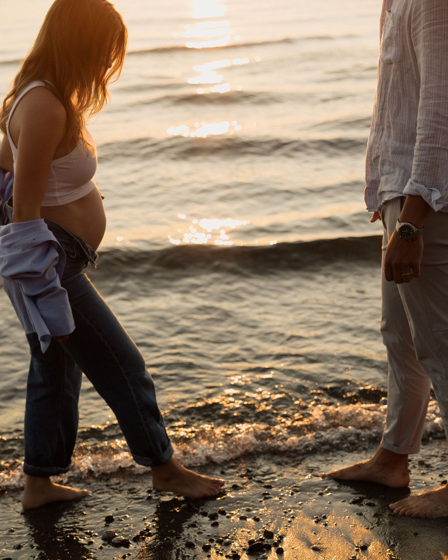 A couple standing barefoot on a beach at sunset with waves gently lapping at their feet.